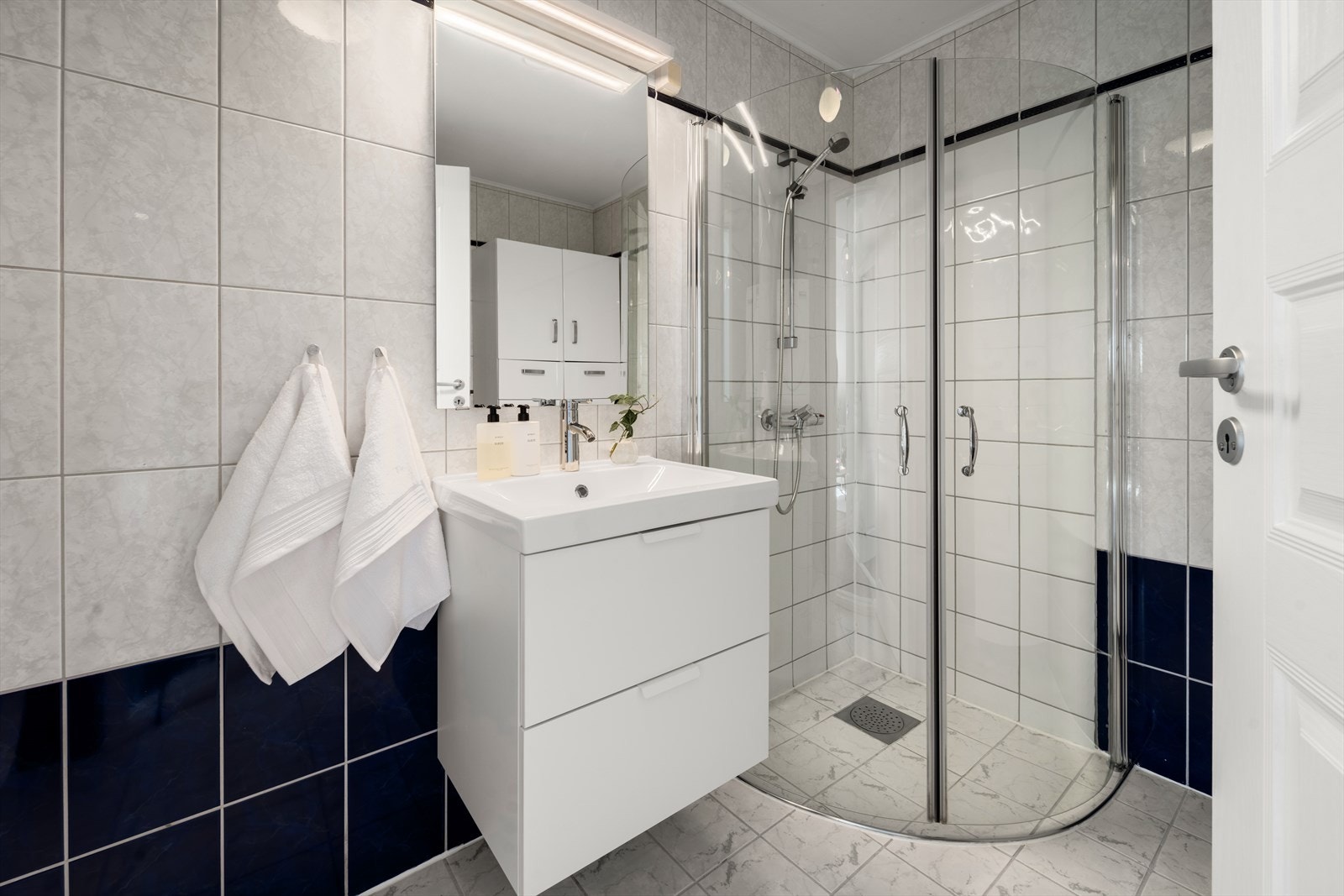 Flislagt baderom bestående av vegghengt servant med underskuffer, WC og dusjsone med innfellbare glassdører Galleribilde