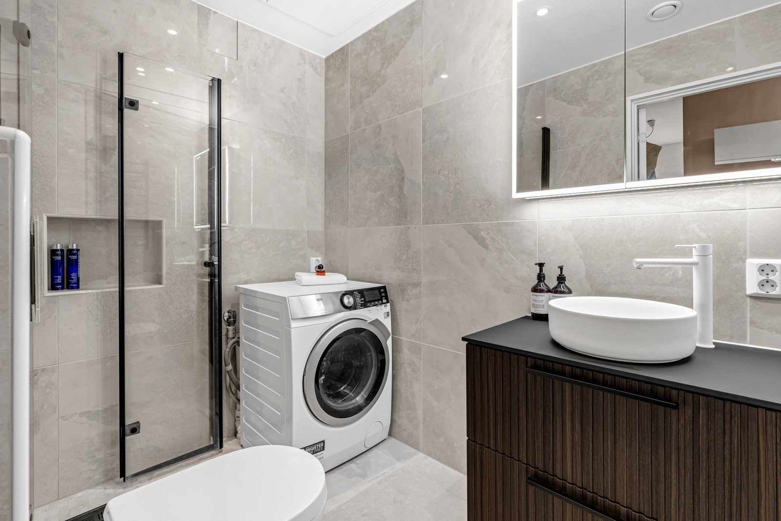 Vegghengt toalett med innebygget sisterne, dusjsone med innfellbare dusjdører, overskap med speilfronter og belysning. Galleribilde