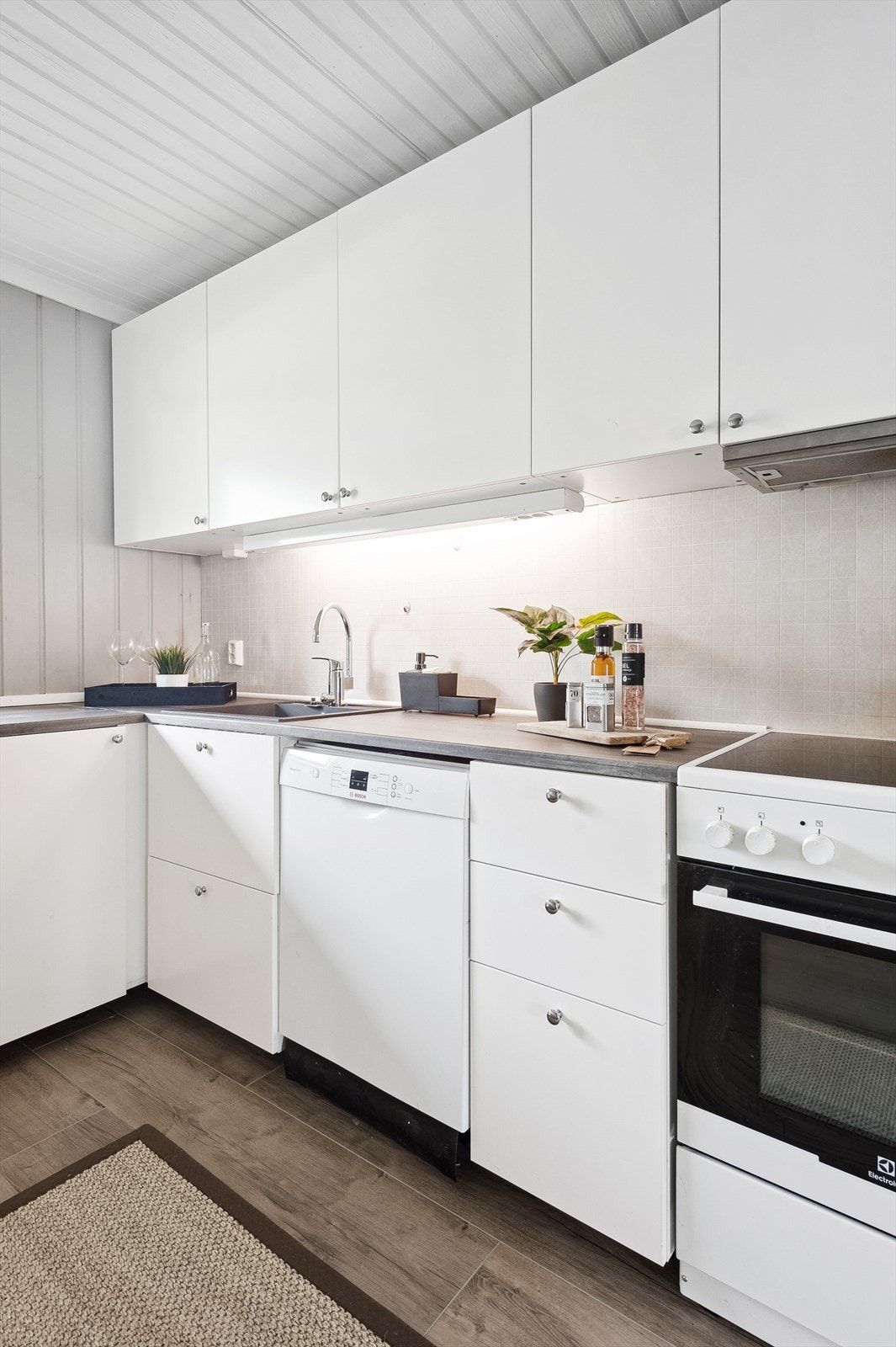 Innredningen har hvite glatte fronter og laminert benkeplate med nedfelt oppvaskkum. Praktisk med flisplate i benkeryggen og belysning under overskapene. Galleribilde