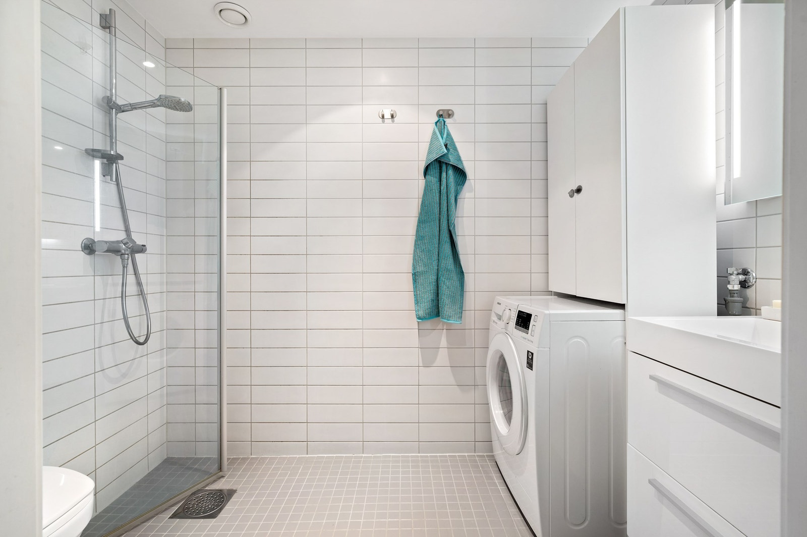 Badet er pent flislagt med lyse fliser og varme i gulvet. Baderomsinnredningen består blant annet av vegghengt servantinnredning med skuffer og ovenpåliggende servant. Galleribilde