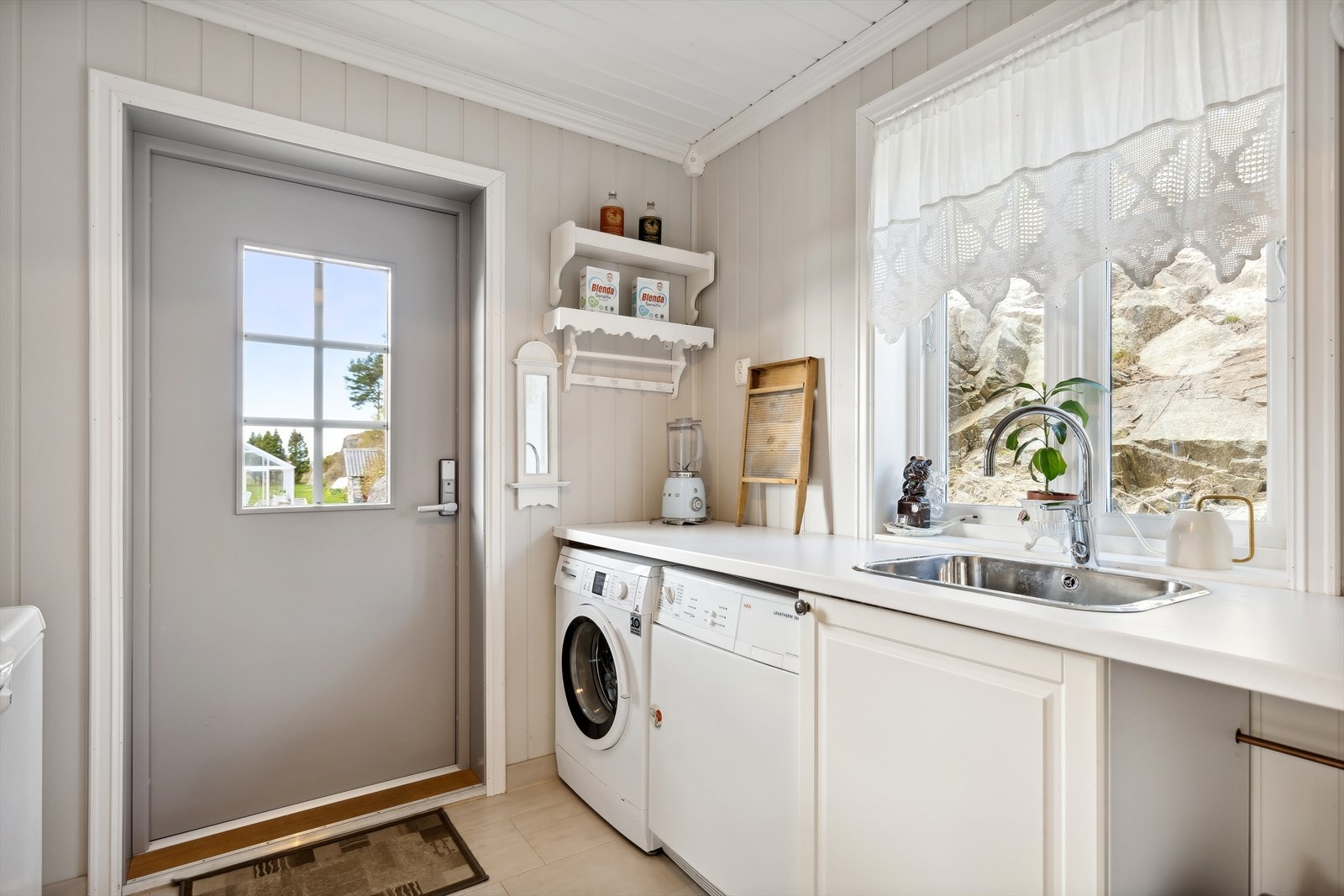 Praktisk vaskerom/biinngang med fliser på gulv og malte panelplater på vegg. Vaskerommet har utslagsvask, opplegg for vaskemaskin og varmtvannsbereder. Galleribilde