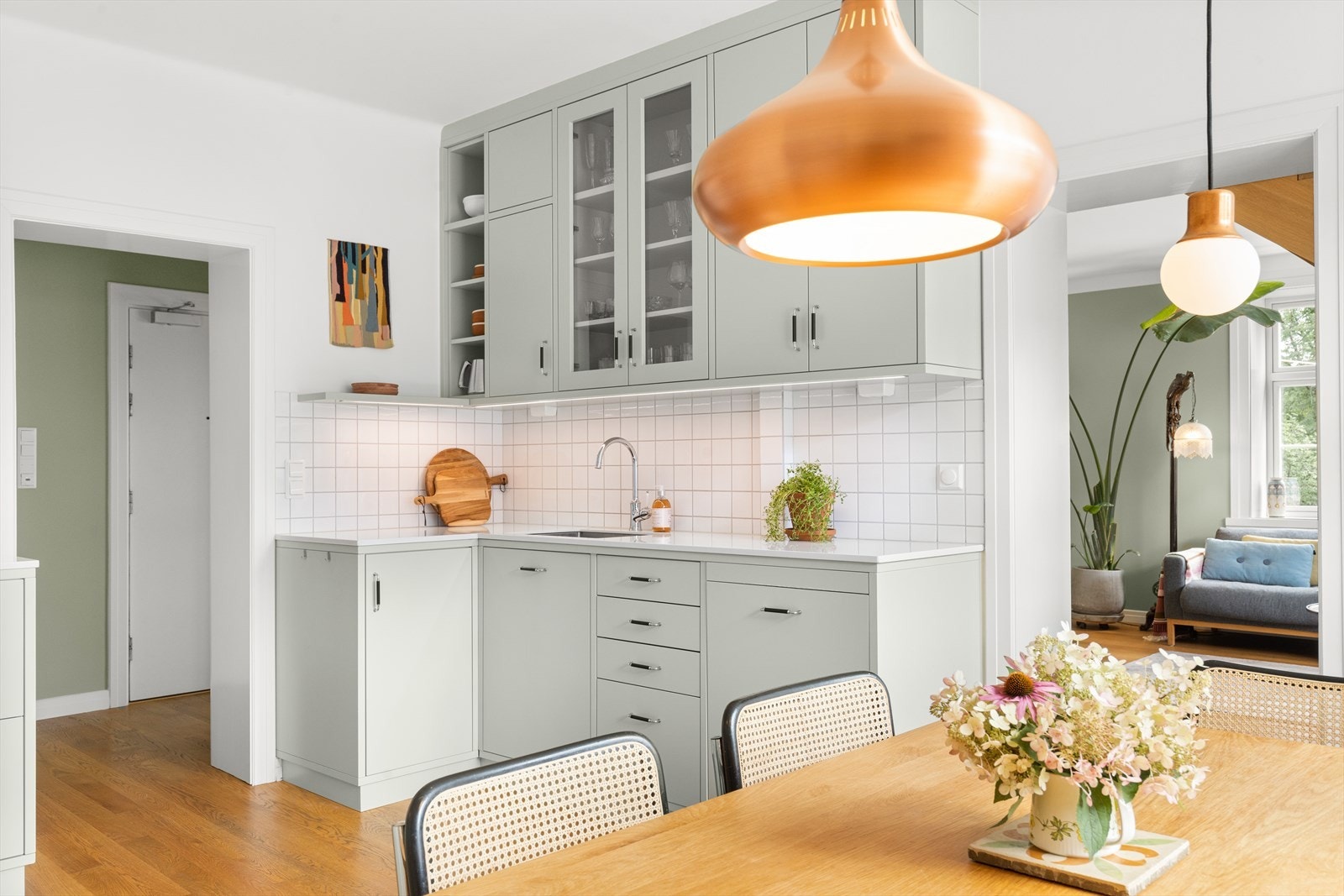 Kjøkkeninnredning bestående av glatte fronter, vakker hvit slitesterk kompositt benkeplate med underlimt vaskekum, samt fliser og belysning over benkeplaten. Denne delen av kjøkkeninnredningen har takhøye overskap. Galleribilde