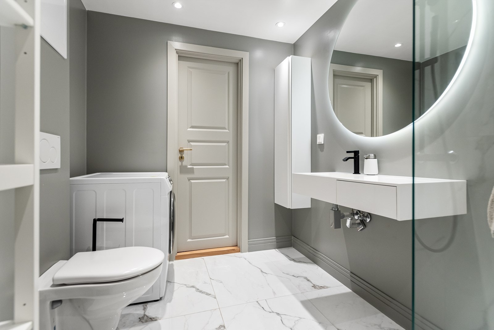 Baderommet er utstyrt med servant med skuff, speil med LED-lys, høyskap på vegg, opplegg for vaskemaskin, dusjhjørne, vegghengt toalett og downlights i himling. Galleribilde