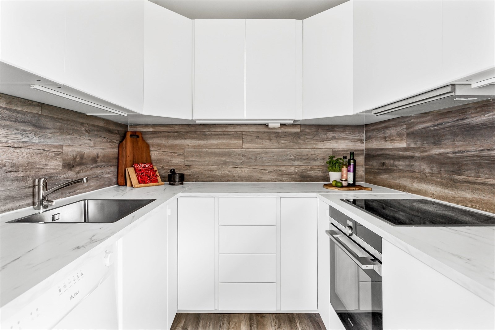 Hvit, glatt innredning med skuffer og skap, lys benkeplate og laminatgulv over benken. Galleribilde