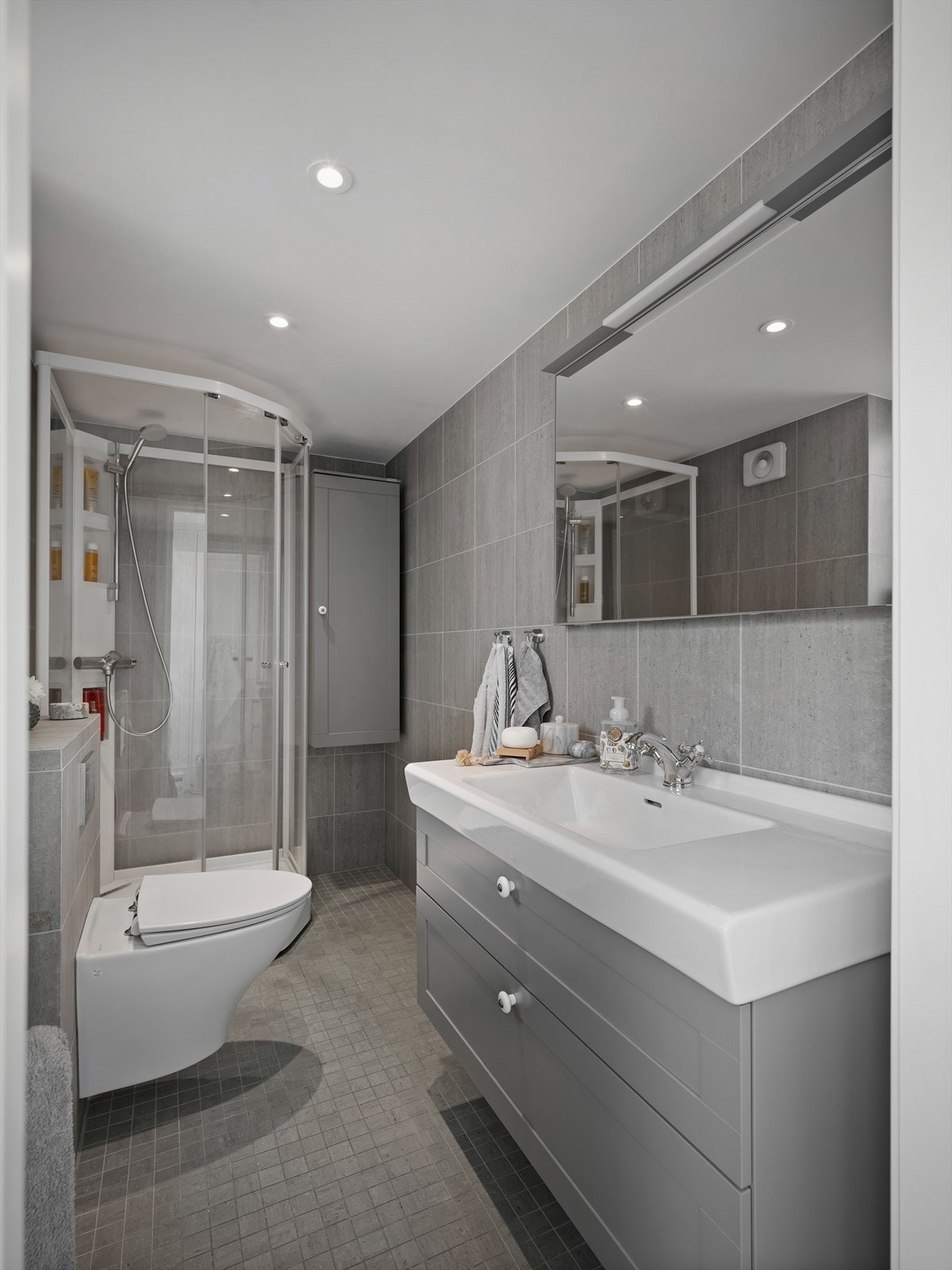 Badet er innredet med et veggmontert toalett, stilren baderomsinnredning med heldekkende servant, speil med overliggende belysning og dusjkabinett. Galleribilde