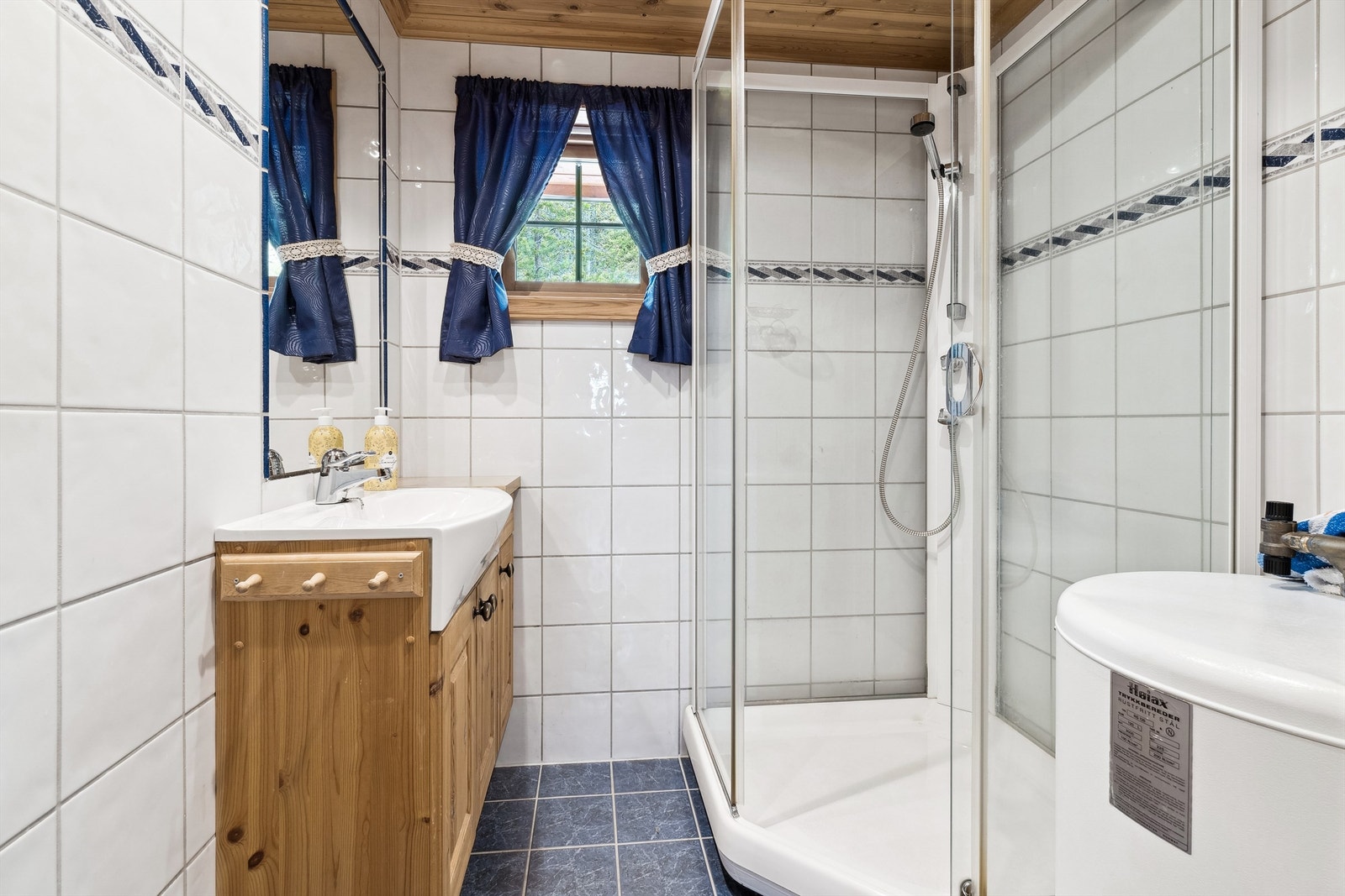 Badet har hvite fliser på veggene og blå fliser på gulvet. Badet er innredet med servant i skapinnredning, dusjkabinett og varmtvannsbereder. Galleribilde