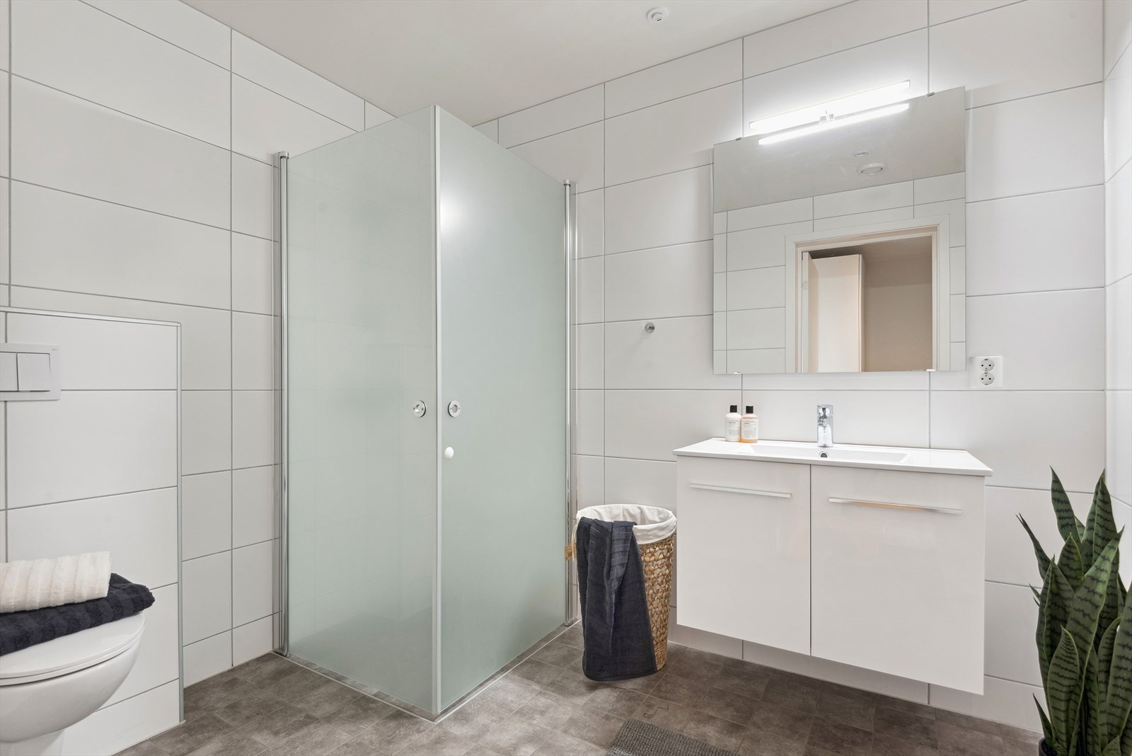 Begge baderommene er utstyrt med vegghengt toalett, servant med underskap og overhengt speil samt dusjvegger. Ventileringen ivaretas av ventilasjonsvifte og tilluft i dør. Galleribilde