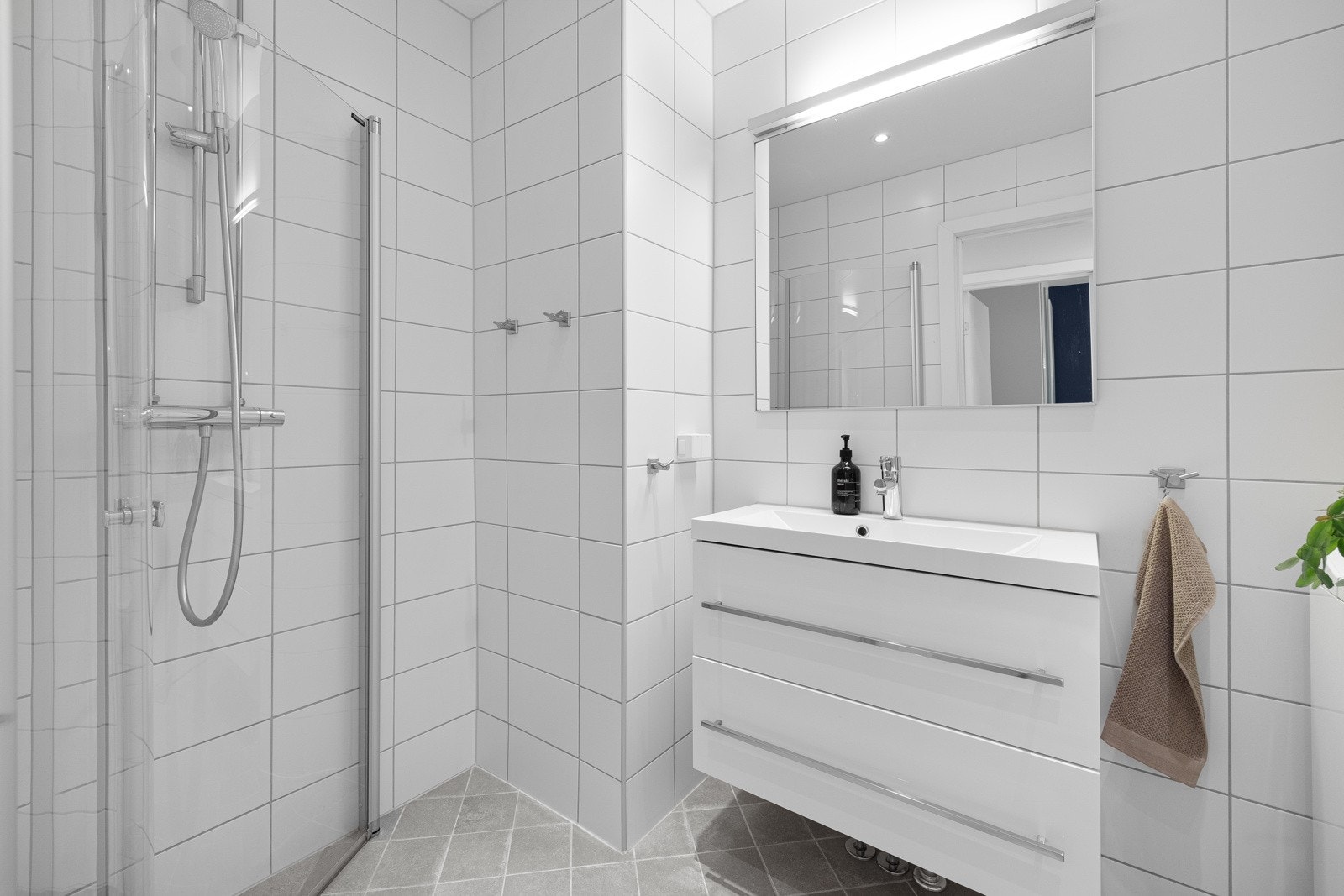 Tidløs innredning med nedfelt servant med skuffer for oppbevaring, speil med belysning over og hjørnedusj med glassdører. Det er opplegg for vaskemaskin på badet. Galleribilde