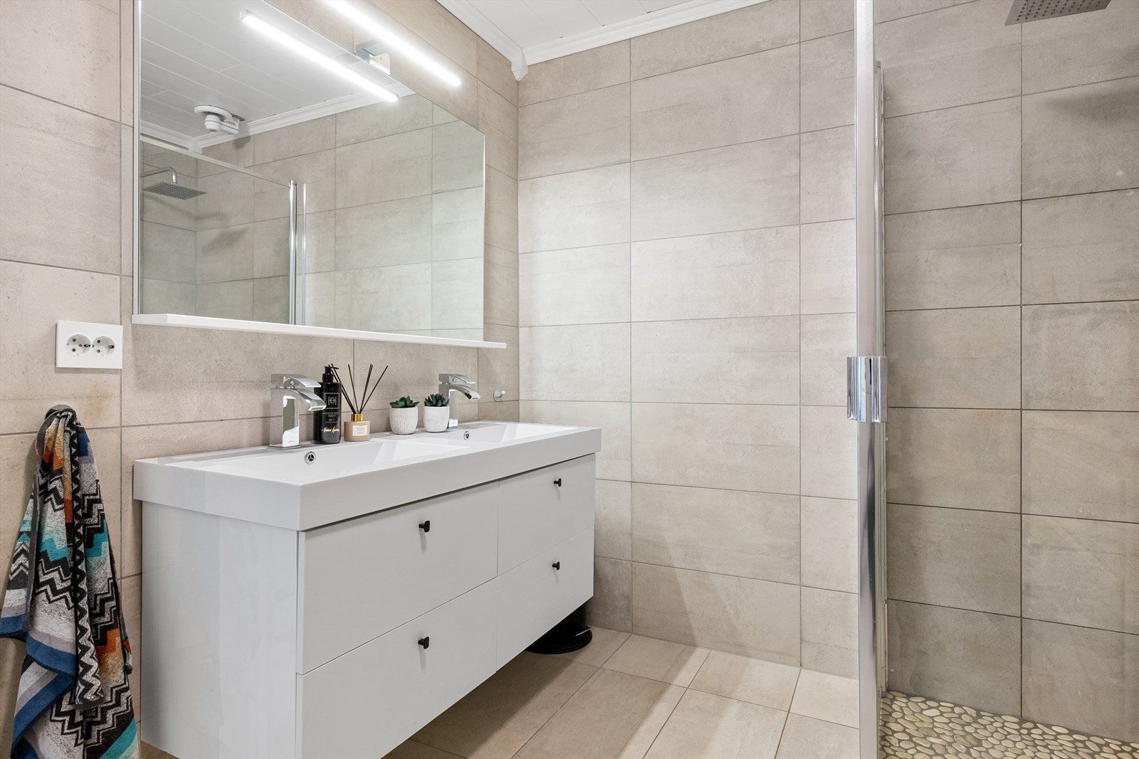 Lekkert bad med fliser på gulv og vegg. Rullestein i dusjsonen. Dobbel vask i seksjon og speil med belysning. Galleribilde