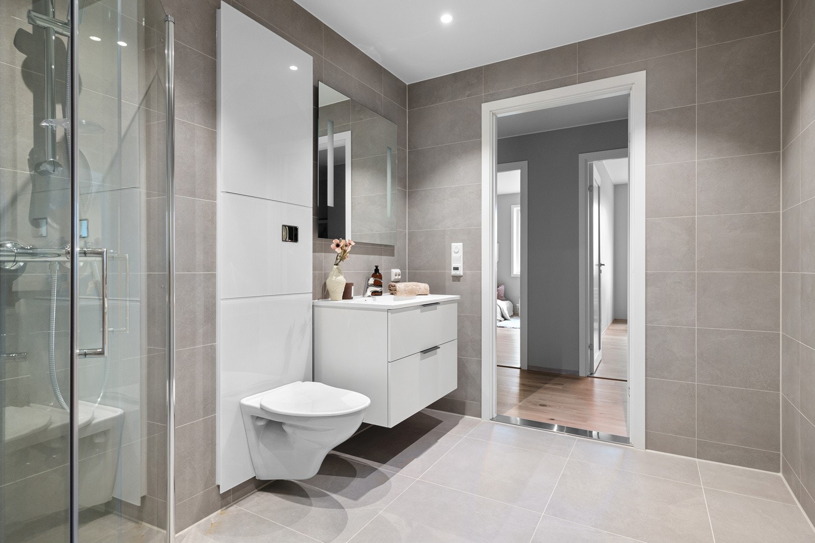 Badet er tidløst og moderne, med lysegrå fliser på gulvet med gulvvarme og mørkegrå fliser på veggflatene. Baderomsinnredningen består av en heldekkende servant på en hvit skuffeseksjon og speil med integrert belysning. Galleribilde