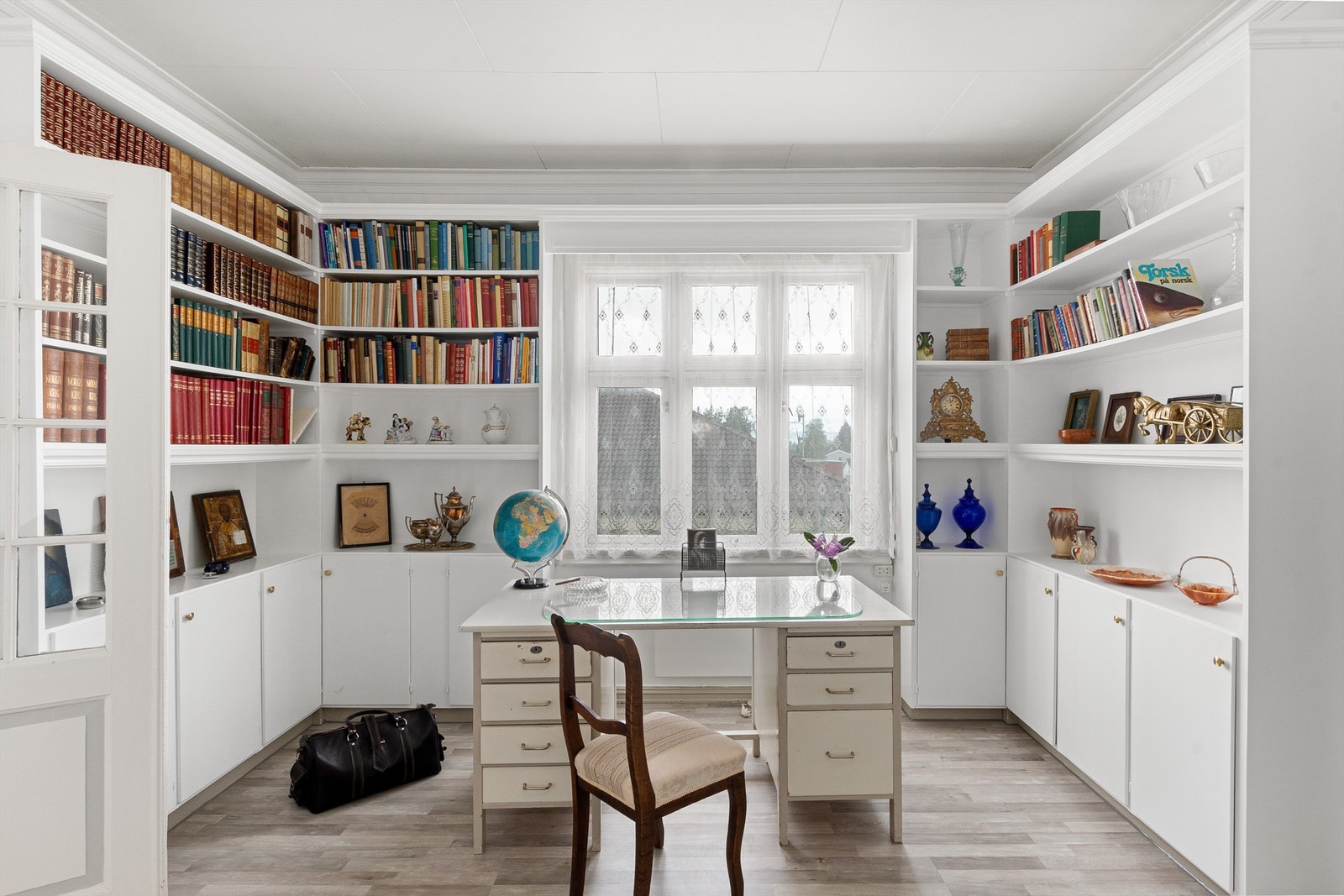 Biblioteket med hvitmalte veger og laminat på gulvet. Rommet er innredet med plassbygde bokhyller med skapdører på nederste del. Her er det mulig med hjemmekontor, gjøre lekser eller man trenger en plass å nyte hobbyen sin. Galleribilde