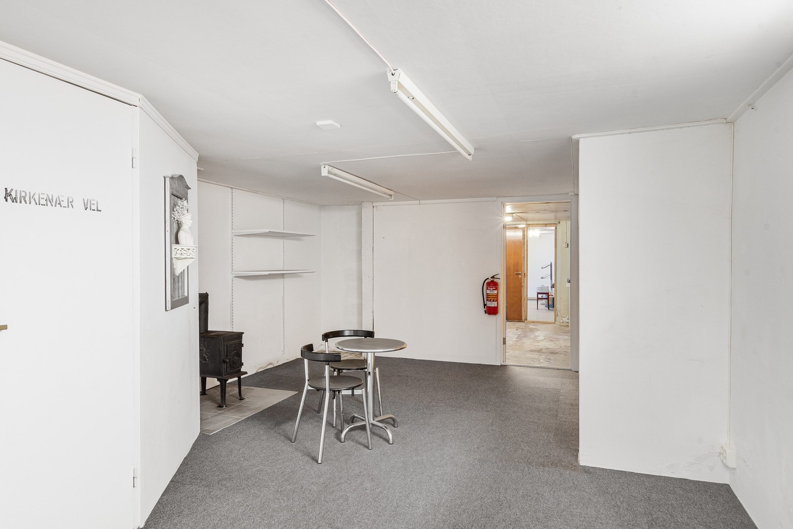 Videre inn kommer man til et kjellerrom med grått teppe på gulvet og hvitmalte vegger. Her er det etablert en vedovn. Galleribilde