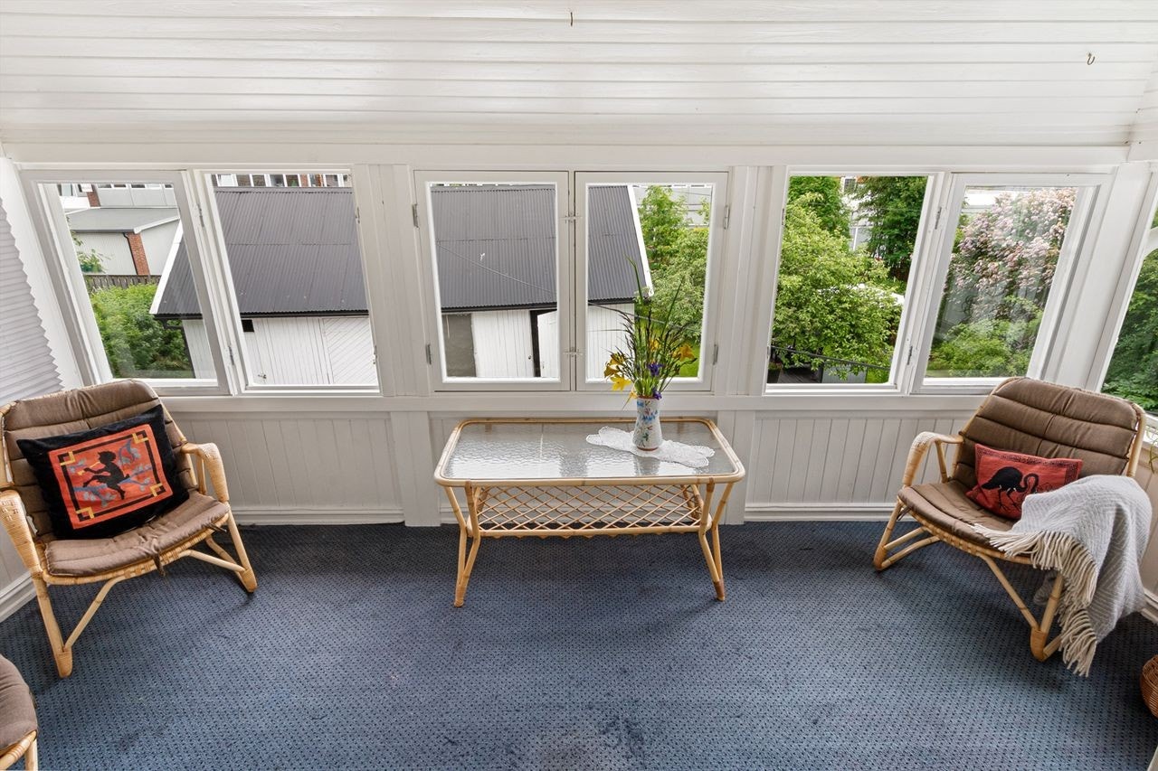 Innglasset balkong med hvitmalt panel på veggene og blått teppe på gulvet. Vinduer på alle veggene gir rommet mye dagslys inn og litt følelsen av å sitte ute selv om man sitter tørt og godt innendørs. Galleribilde