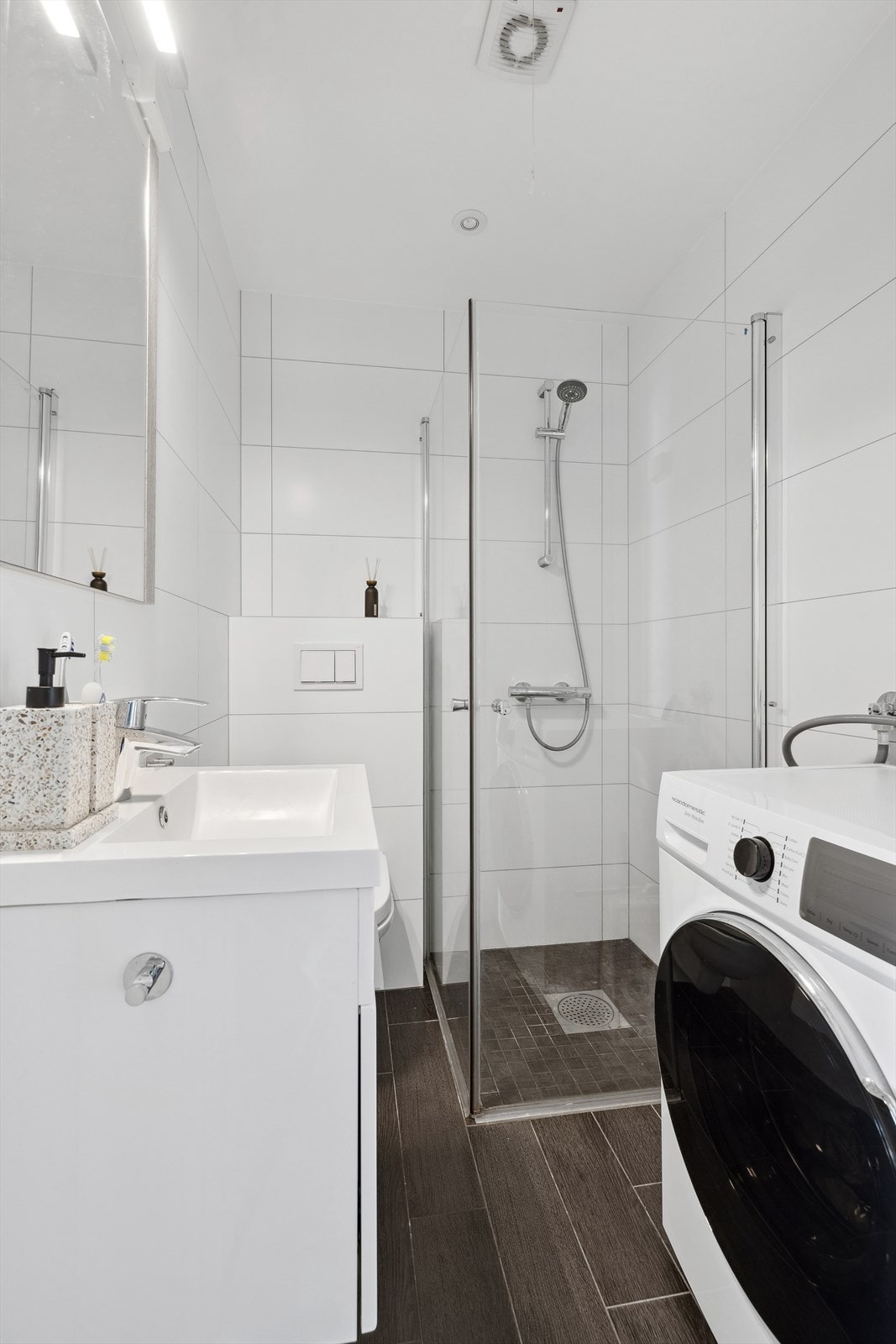 Badet er utstyrt med dusjhjørne med innfellbare glassvegger, vegghengt wc, servantinnredning med skuff, samt opplegg og plass til vaskemaskin Galleribilde