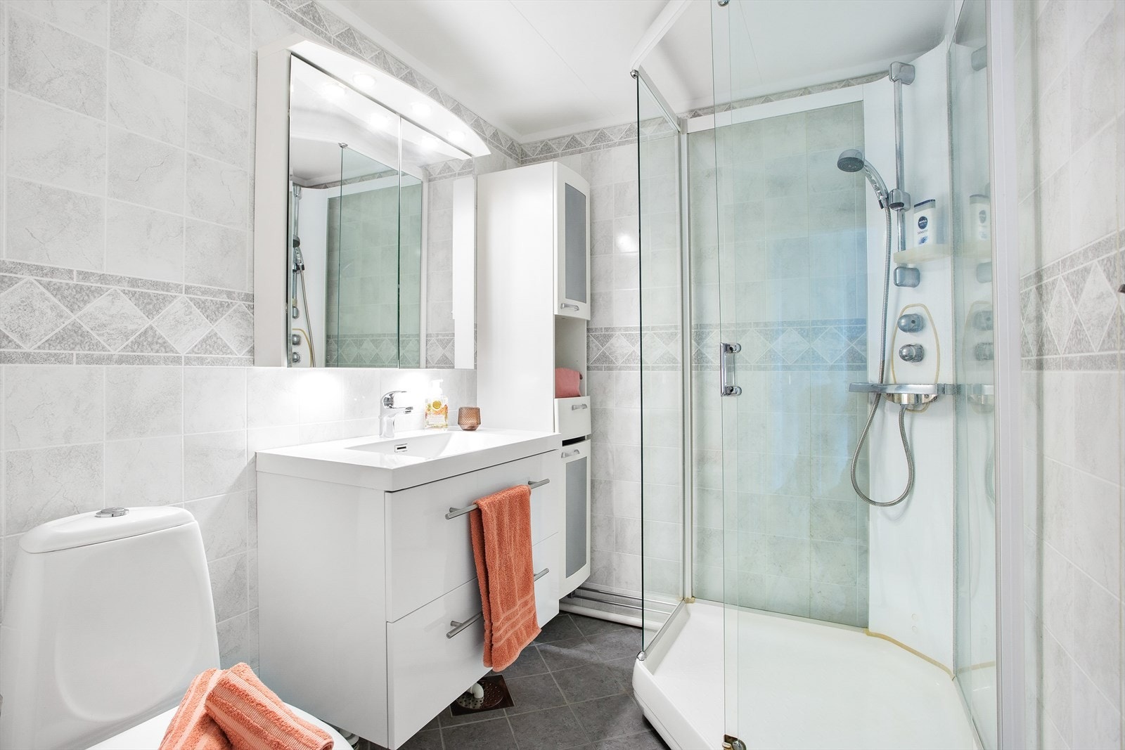 Rommet er praktisk innredet med heldekkende servant på hvit skuffeseksjon, veggskap med frostede dører, speil med overlys, et toalett og et dusjkabinett. Galleribilde