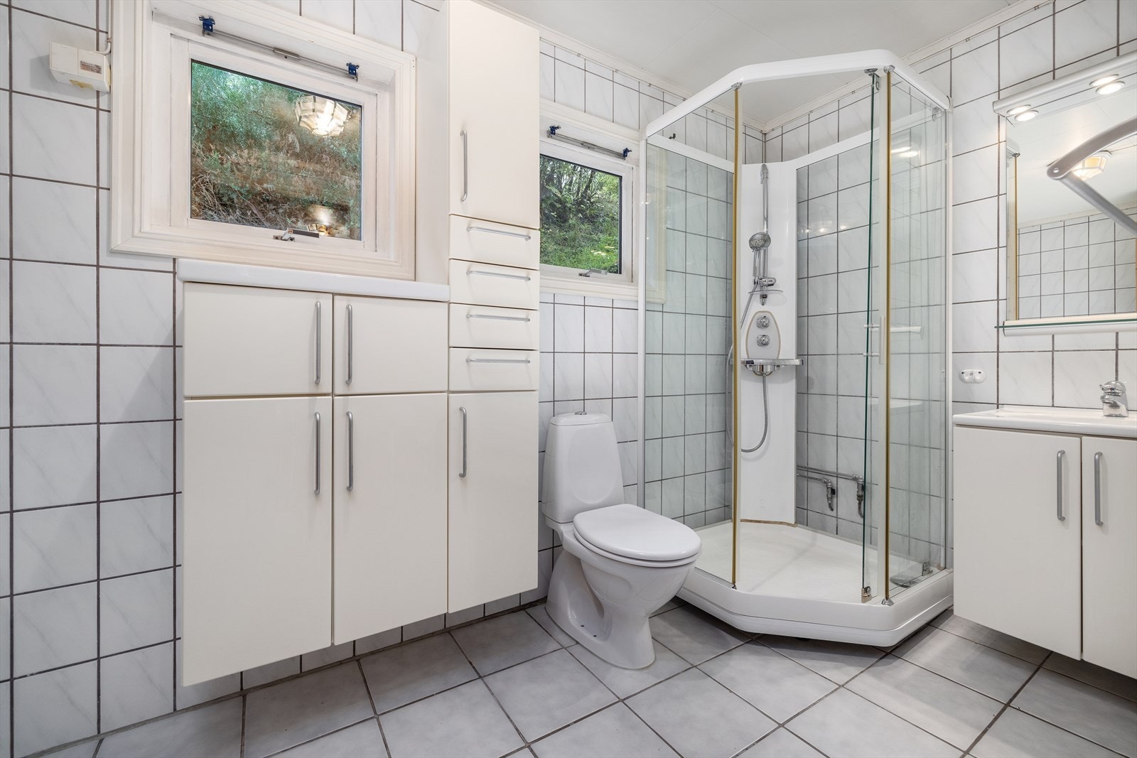 Badet har fliser på gulv/vegger og er innredet med servant. dusjkabinett, gulvstående toalett, skyllekum og opplegg for vaskemaskin. Galleribilde