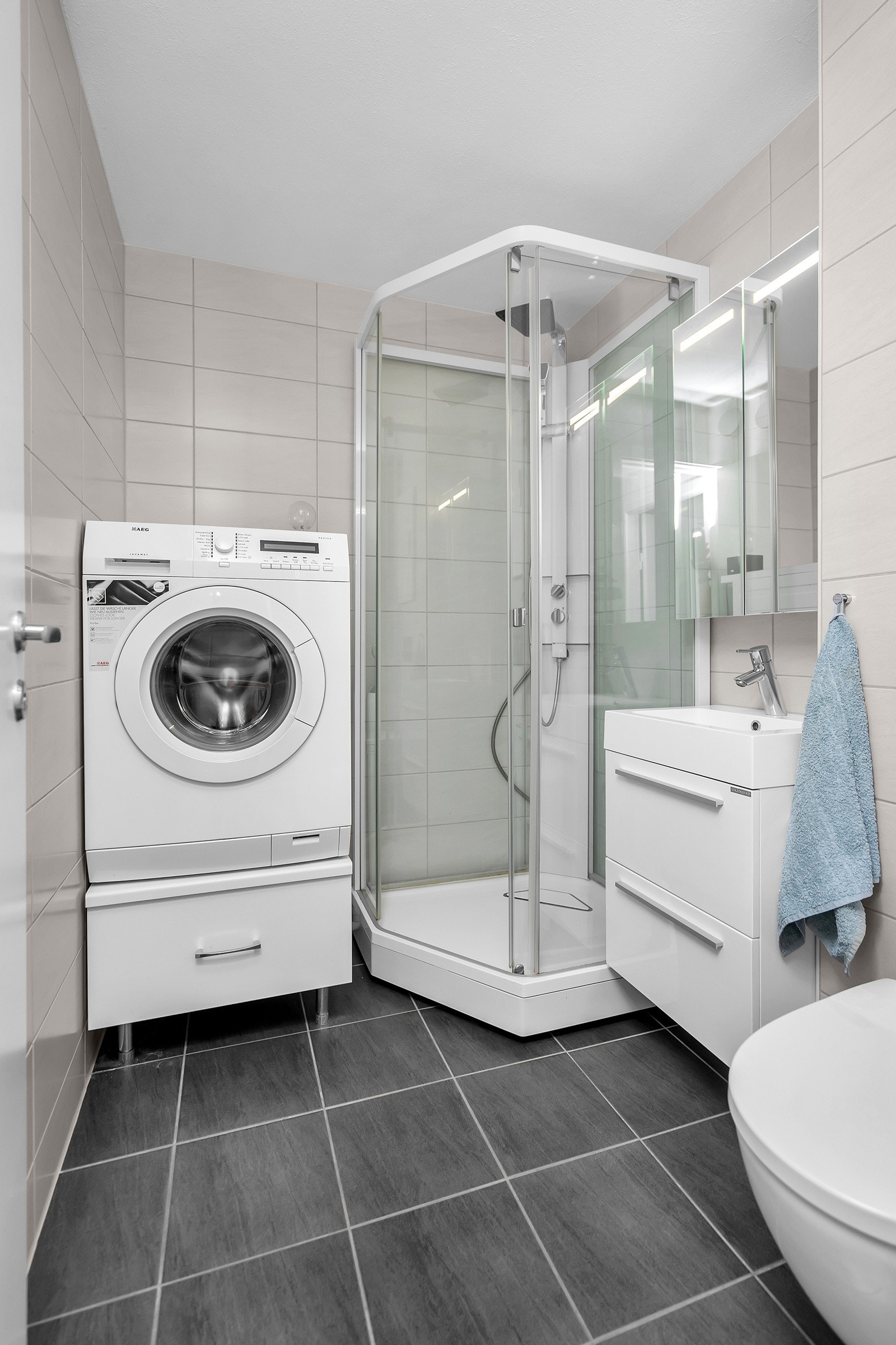 Flott flislagt bad med dusjkabinett, servant med innredning, vegghengt toalett og opplegg til vaskemaskin Galleribilde