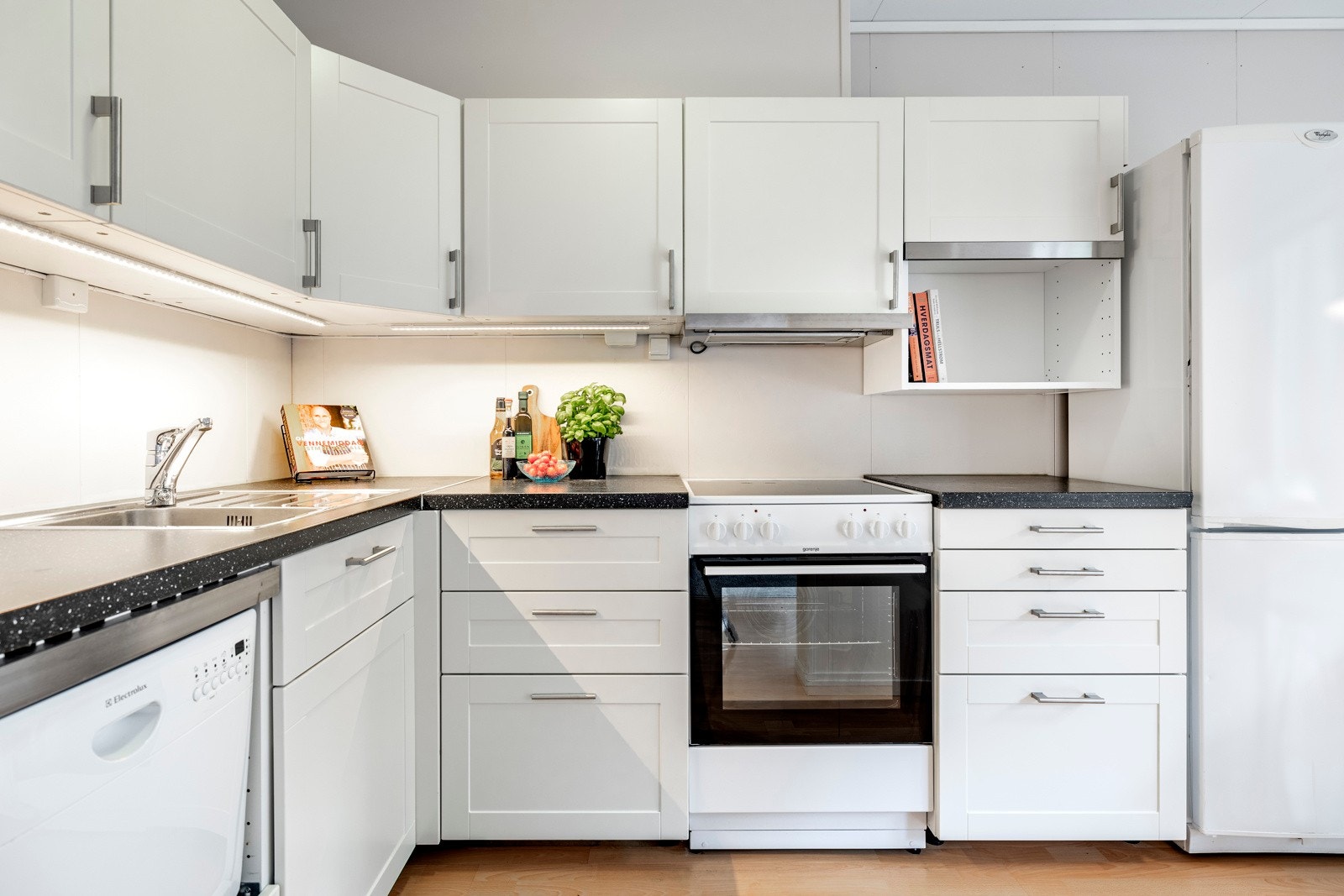 Kjøkkenet har en pen innredning fra Ikea plassert i L-form, åpent mot stuen. Innredningen er holdt i en klassisk stil med hvite, profilerte fronter og benkeplate i mørk laminat med nedfelt vaskekum i stål. Galleribilde