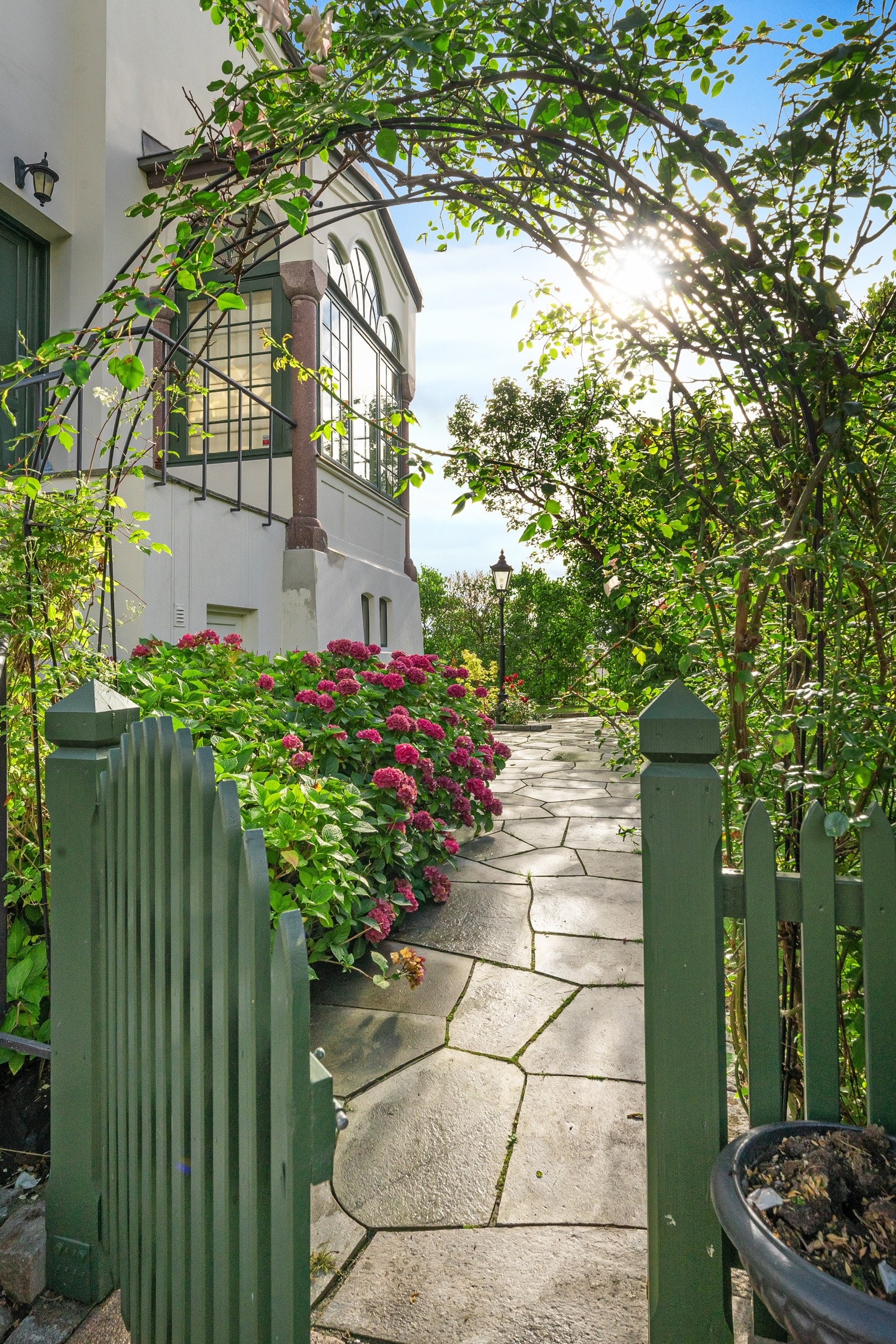 Hagegangen ender i en grønnmalt port med blomsterportal mot gårdsplassen, og omkranses av plen kantet med granittstein og bugnende blomsterbed med hortensia, roser og andre prydvekster Galleribilde