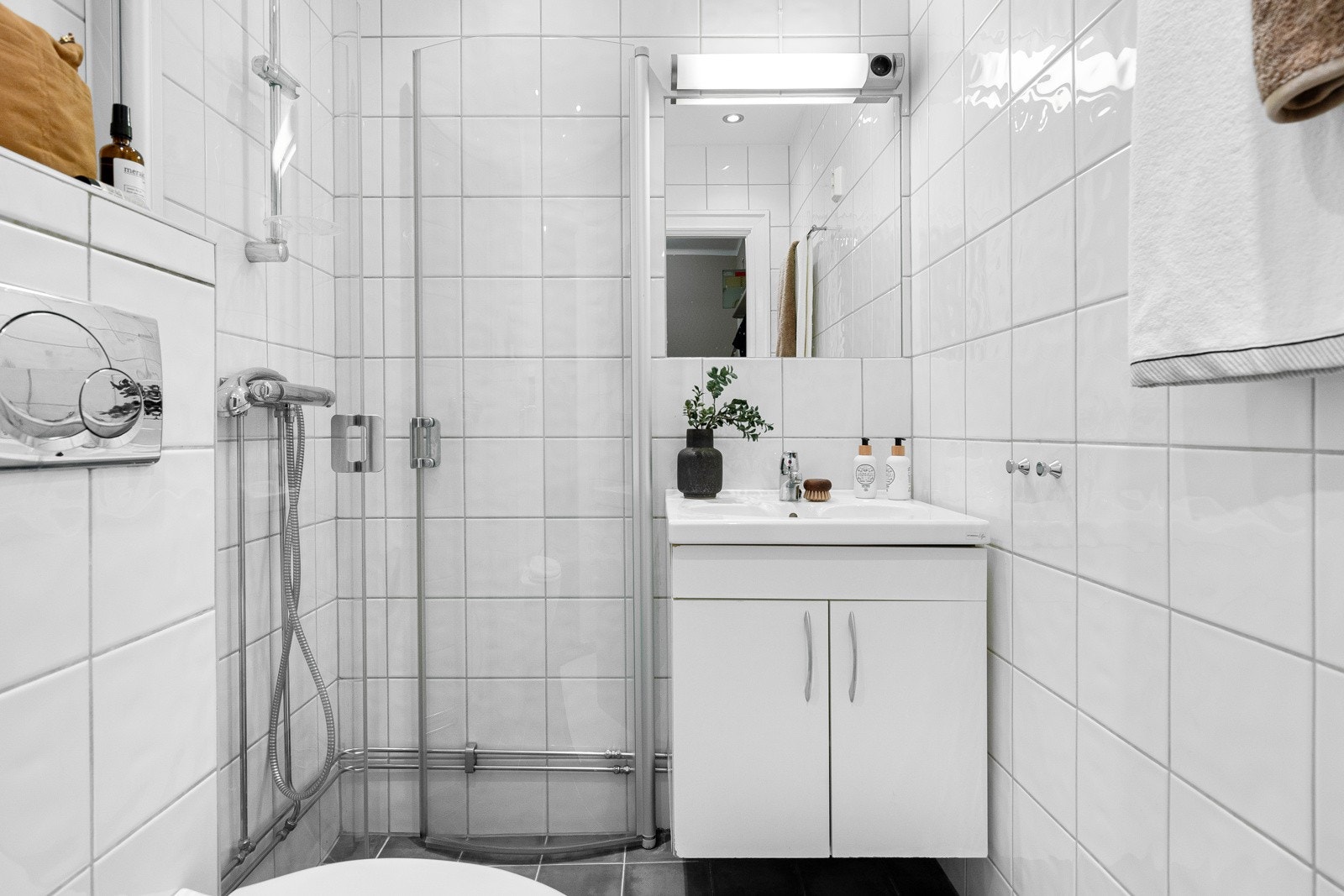 Flislagt baderom som inneholder dusjhjørne med innfellbare dører i glass, benkeskap med heldekkende servant, innfliset speil, lys og toalett. Galleribilde