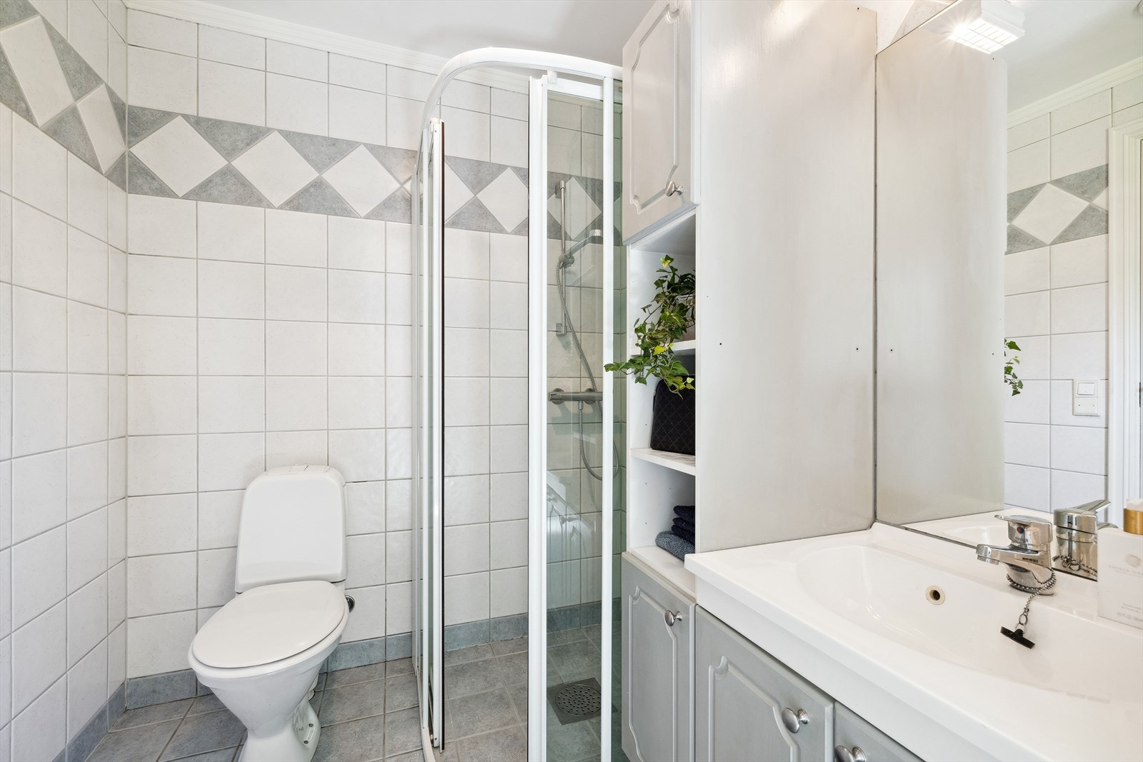 Kombinert bad/wc og vaskerom i 1. etasje. Det er flislagte overflater og malte plater i himling. Baderomsdelen er utstyrt m/dusjhjørne, klosett, hel nedfelt servant og baderomsinnredning med servantskap, høyskap, speil og lys over speilet. Galleribilde