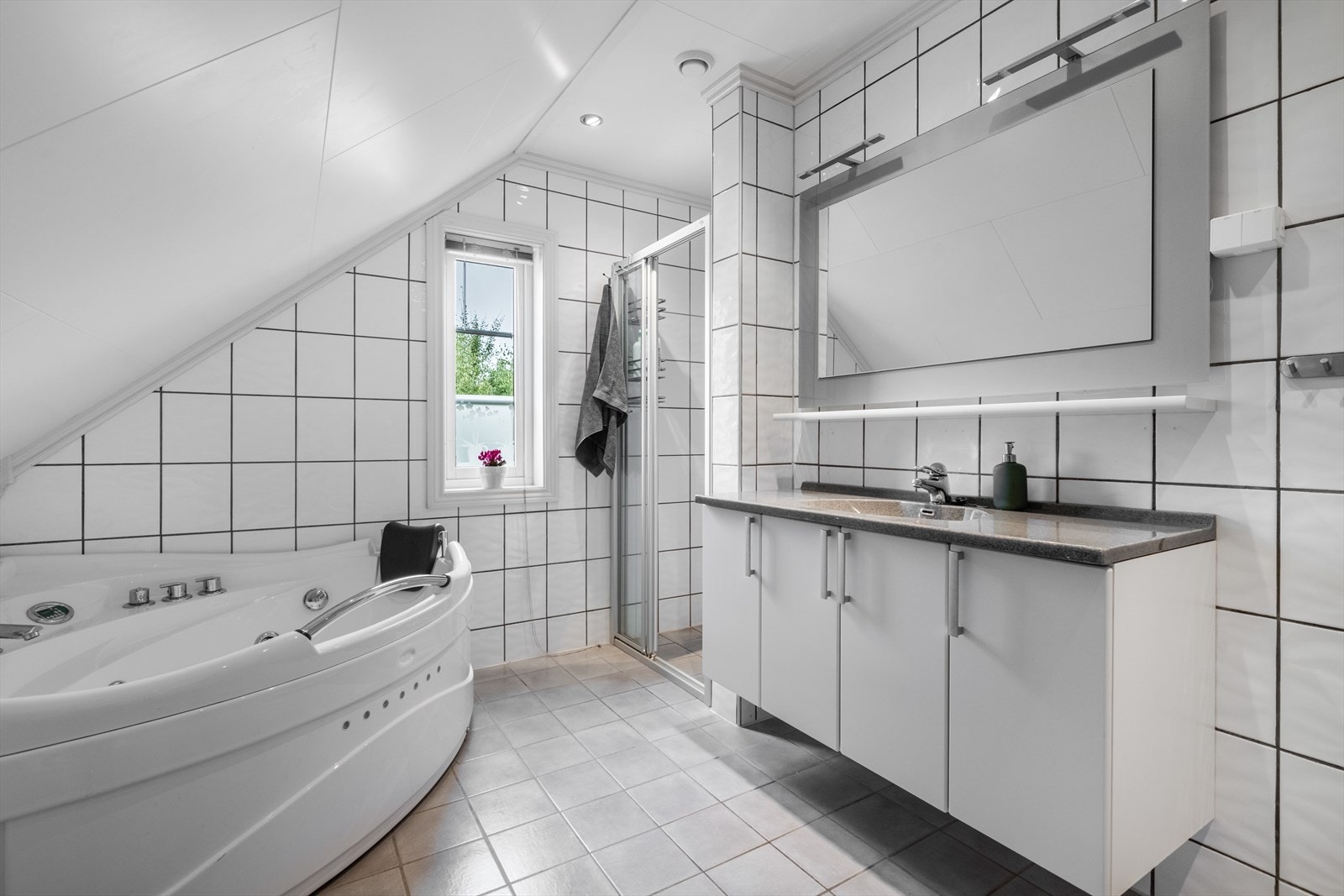 Her er det også et pent bad med flis på gulv og vegger. Badet inneholder toalett, boblebadekar, dusjnisje med skyvedør og innredning. Innredningen har vask med heldekkende benkeplate, underskap med slette fronter og speil med lys. Galleribilde