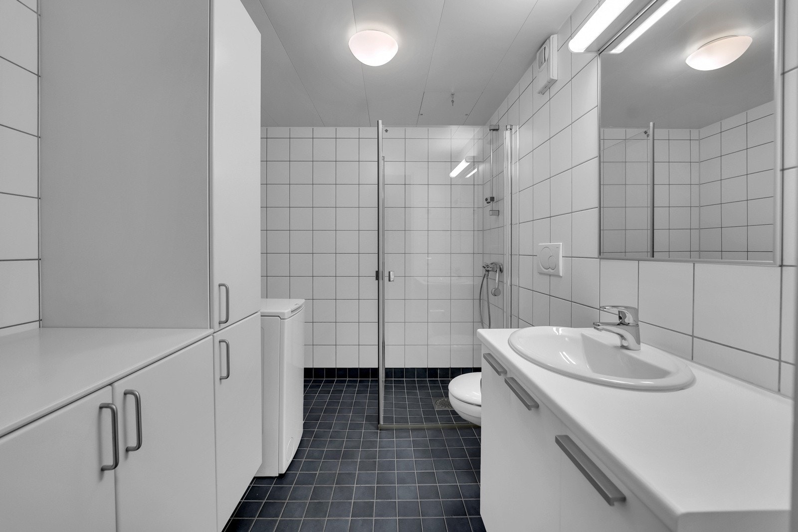 Badet har god størrelse og et delikat uttrykk der mørke gulvfliser kombineres med hvite fliser på veggene. Videre er det vegghengt wc, servant og rikelig med oppbefaring i skap, dusjhjørne med innfellbare dører og opplegg for vaskemaskin. Galleribilde
