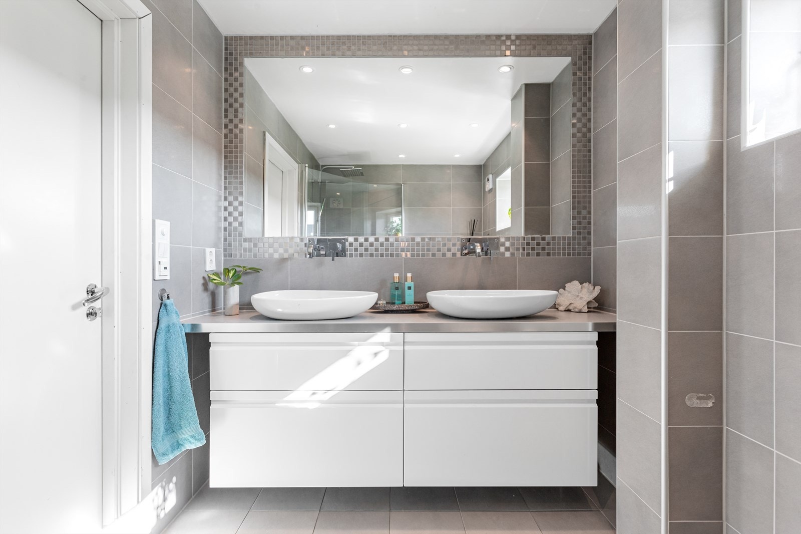 Badet har elektriske varmekabler og elstyrt vifte. Innredningen består av et underskap, 2 servanter, speil, stikk, vannklosett og dusjsone. Galleribilde