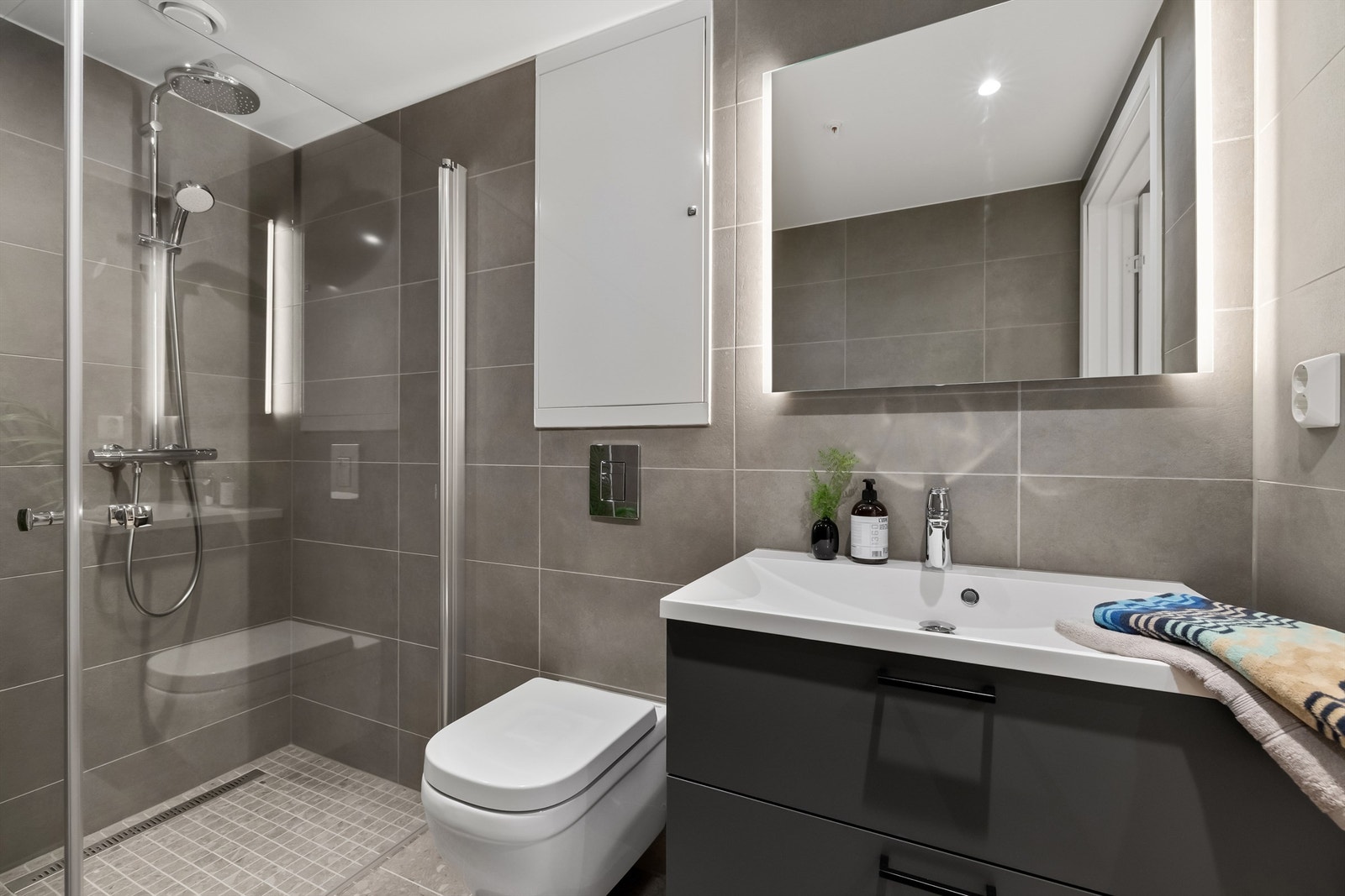 Utenpåliggende speil med innfelt LED-lys. Dusjvegger (rette, svingbare) i herdet glass med aluminiumsprofil. Vegghengt toalett med innebygget sisterne, Grohe Euro (Soft-close toalettsete). Galleribilde