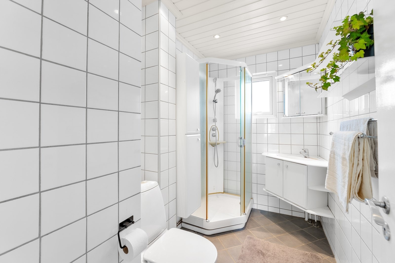 Tidløst baderom med flislagte gulv og vegger. Badet er utstyrt med toalett, servant med skap og dusjkabinett. Galleribilde