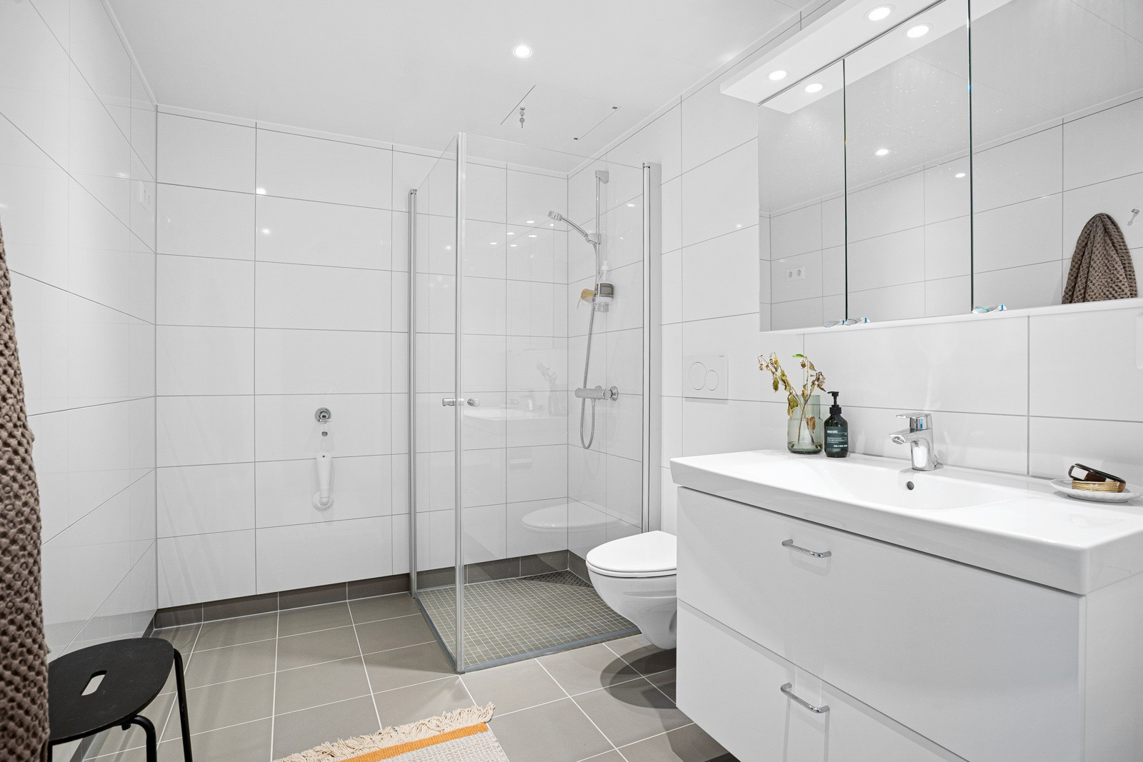 Flislagt hovedbad med praktisk innredning, dusj med innfellbare glassdører og vegghengt toalett.