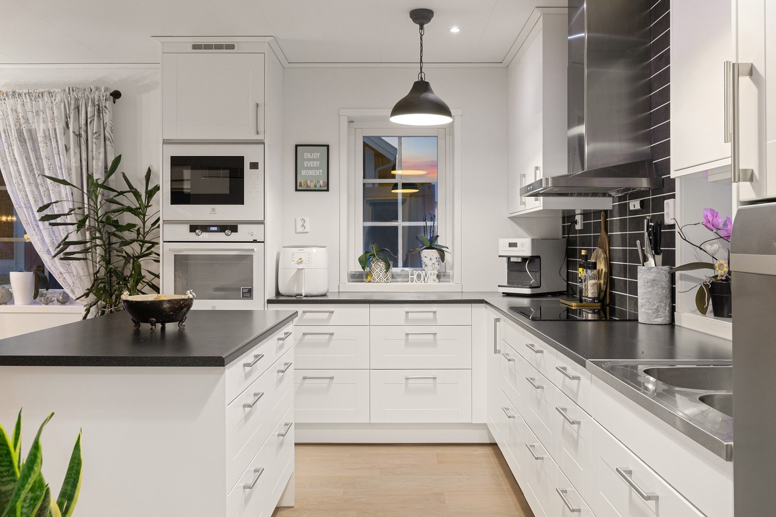 Kjøkkeninnredning med hvite slette fronter, laminert benkeplate med nedfelt platetopp og dobbel vask med ett-greps blandebatteri. Over kjøkkenbenken er det våtromsplater. Galleribilde