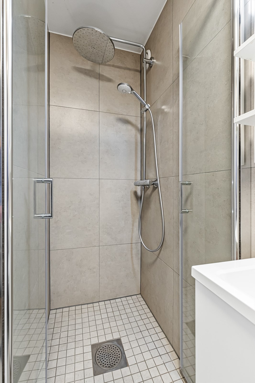 Dusjsonen er hevet 10 cm mot resten av badet og består av glassdører og vegghengt dusjarmatur med regndusj. Badet er ellers innredet med vegghengt servantinnredning med skap, vegghengt speil med lys og hyller og vegghengt toalett. Galleribilde