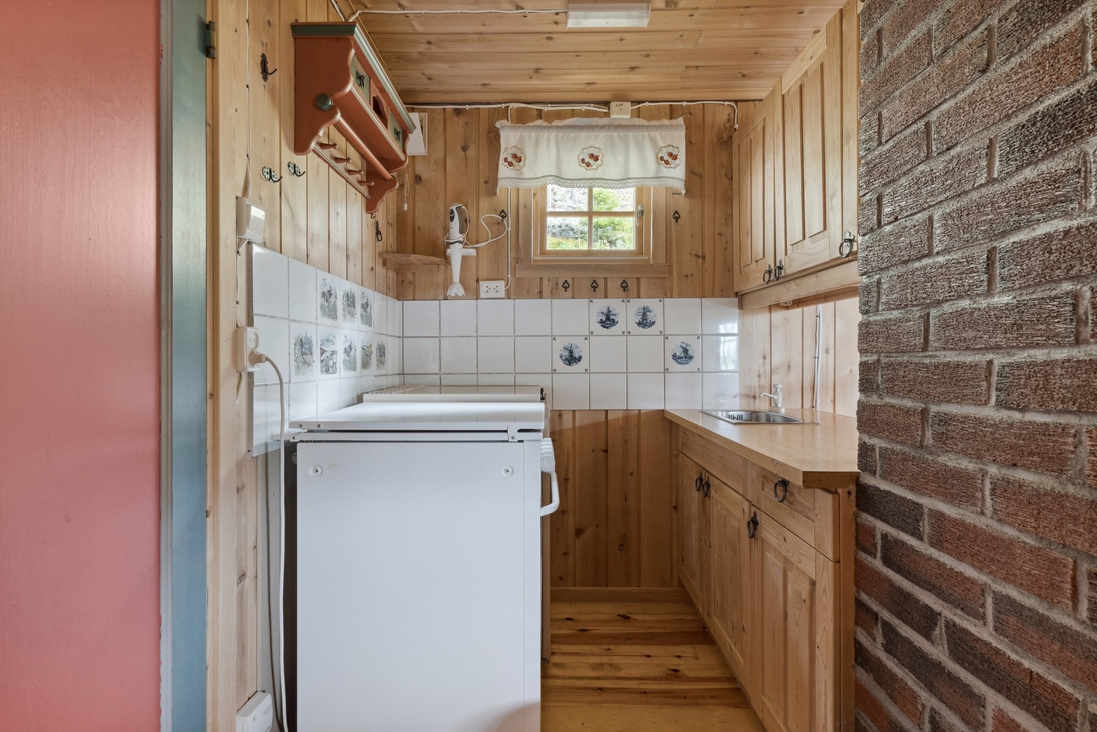 Det er etablert vaskekum med avløp ut i grunnen via rør gjennom gulvet. Det er etablert kjøleskap og komfyr som går på propan. Barløsning mot stuen. Hvite fliser, noen med blått mønster på veggflatene over kjøkkenbenker. Galleribilde