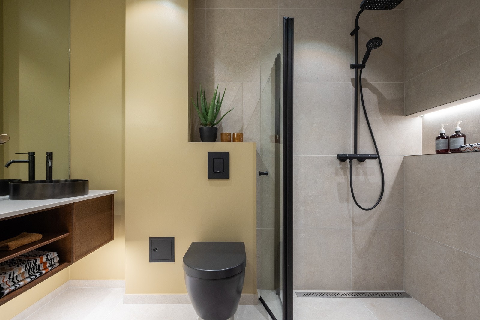 Stort velværebad fra 2024 med varme i gulvstøp, store grå fliser på gulv (60x60) og vegger i dusjsonen. Gultonet vegg med innebygget sisterne over vegghengt klosett. Gulv har sveisemembran og dusjvegger med smøremembran. Galleribilde