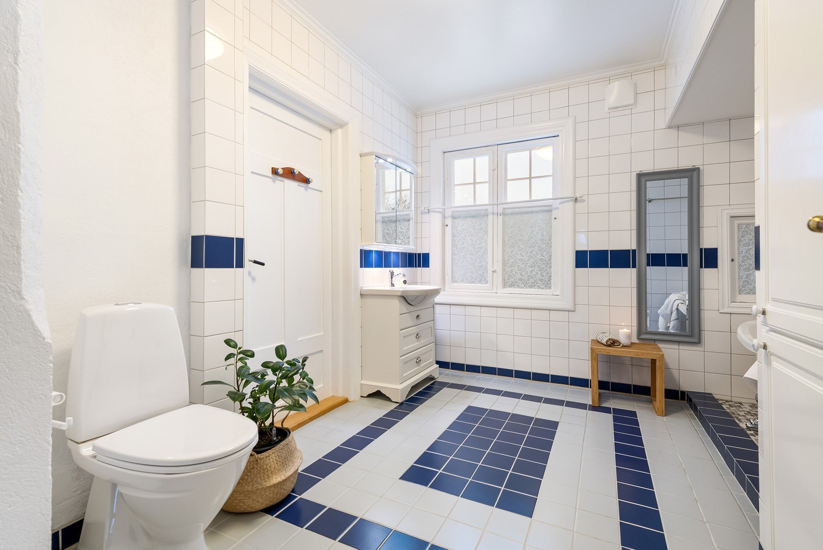 Baderom med hvite og blå fliser på vegger og på gulvet. Baderommet er innredet med servant i lys skuffeinnredning, badekar, dusjnisje med skyvedør og gulvmontert toalett. Galleribilde