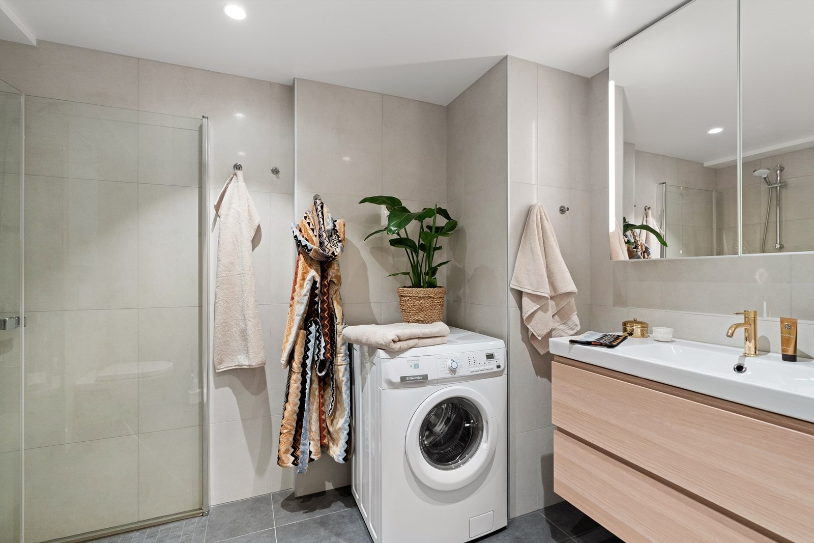 Badet inneholder vegghengt toalett, helstøpt servant, skuffer under servant med slette fronter, skap over med speilfront og integrert lys, dusjdører i klart glass og fordelerskap til rørsystemet Galleribilde