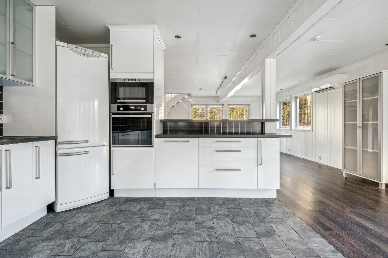 Kjøkken med grått belegg på gulvet ved innredningen og laminat på resten av gulvet. Hvit Ikea innredning med glatte fronter, takhøye skap og laminerte benkeplater. Veggflatene over kjøkkenbenkene med svarte fliser. Galleribilde