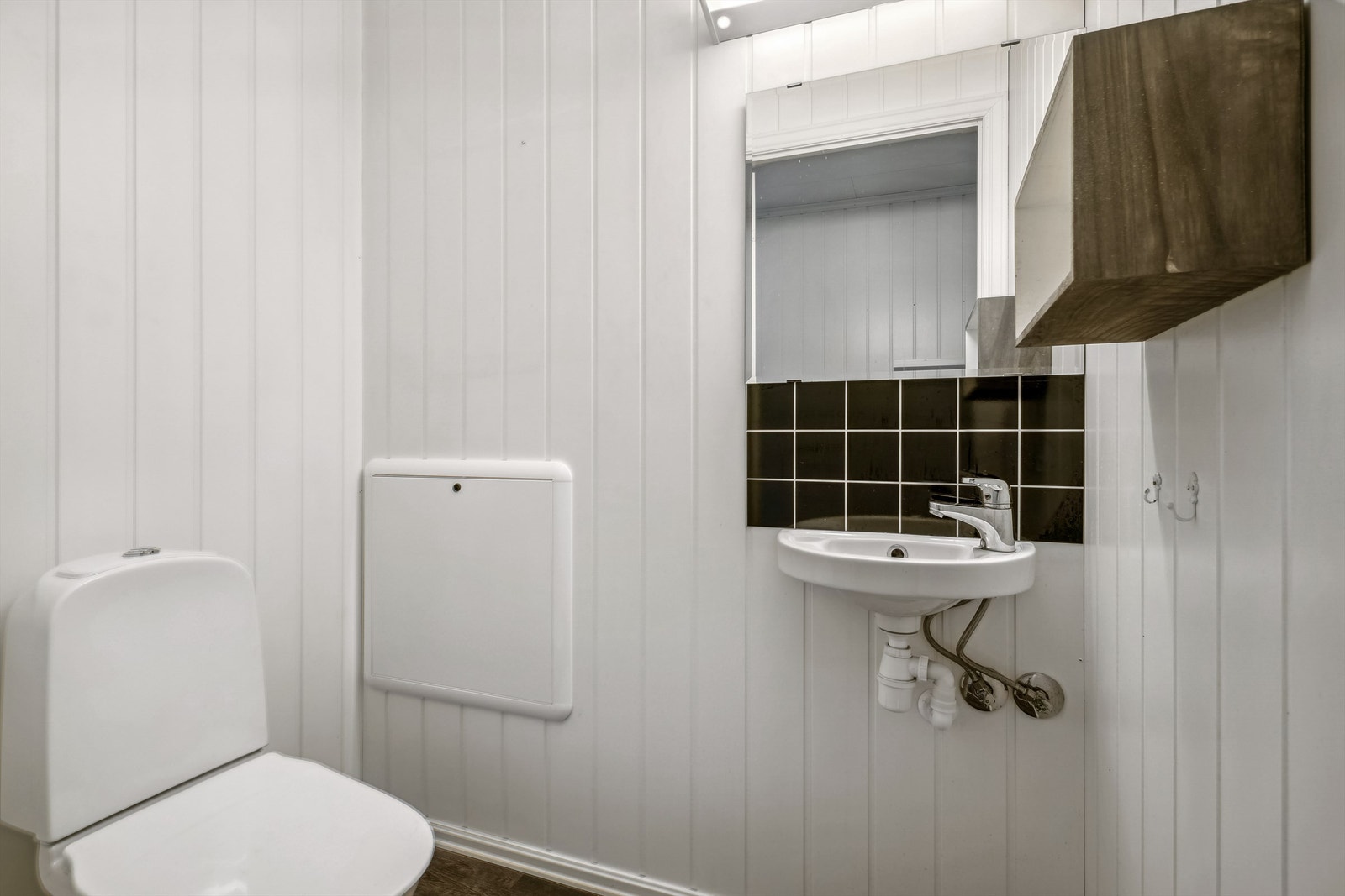 Toalettrom i 1. etasje med hvitmalte panelplater på veggene og laminat på gulvet. Toalettrommet er innredet med servant montert på vegg, veggmontert hylle og gulvmontert toalett. Galleribilde