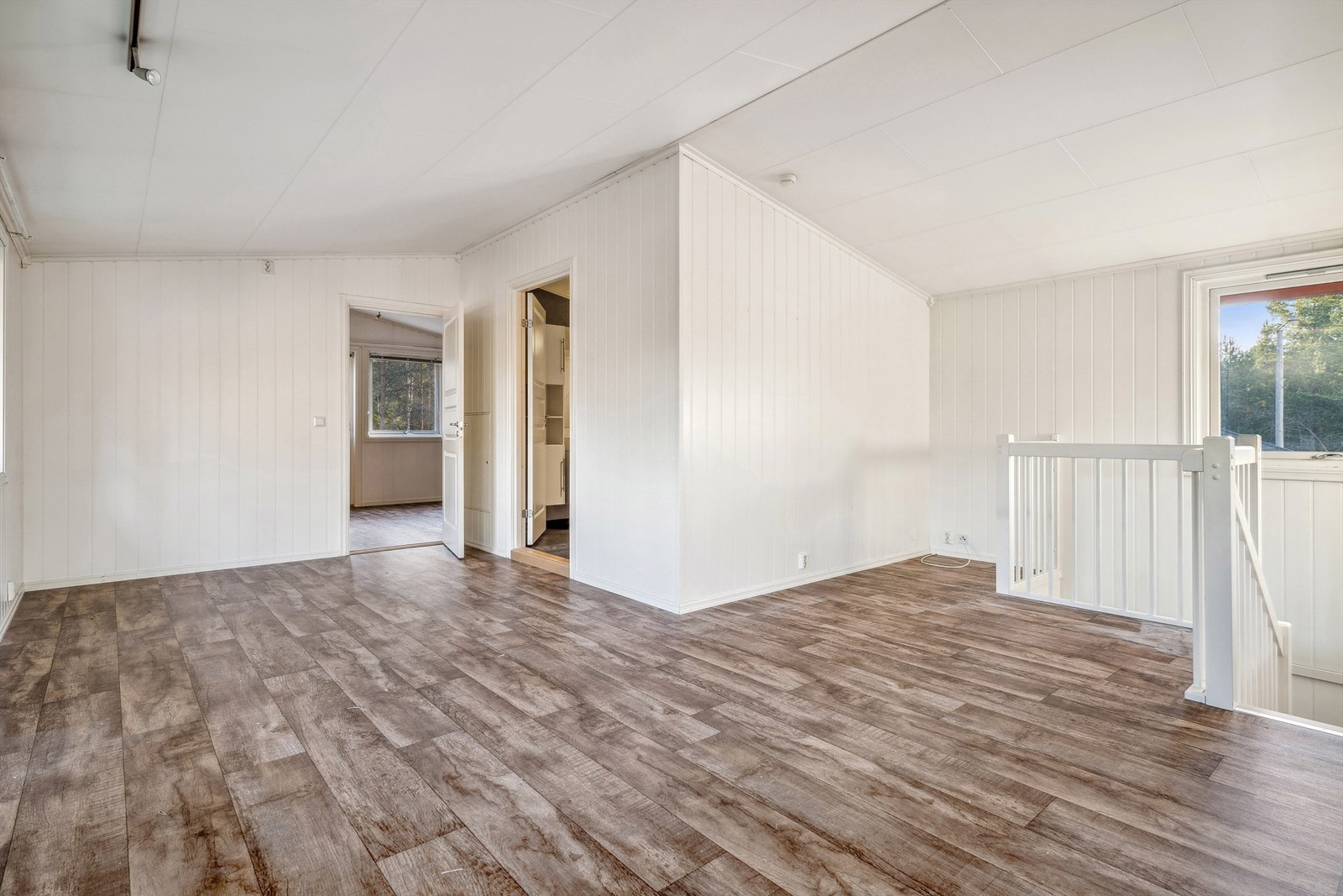 Loftstue med lysmalte panelplater på veggene og brun laminat på gulvet. Rommet er romslig med god plass til sofagruppe og annet møblement. Galleribilde