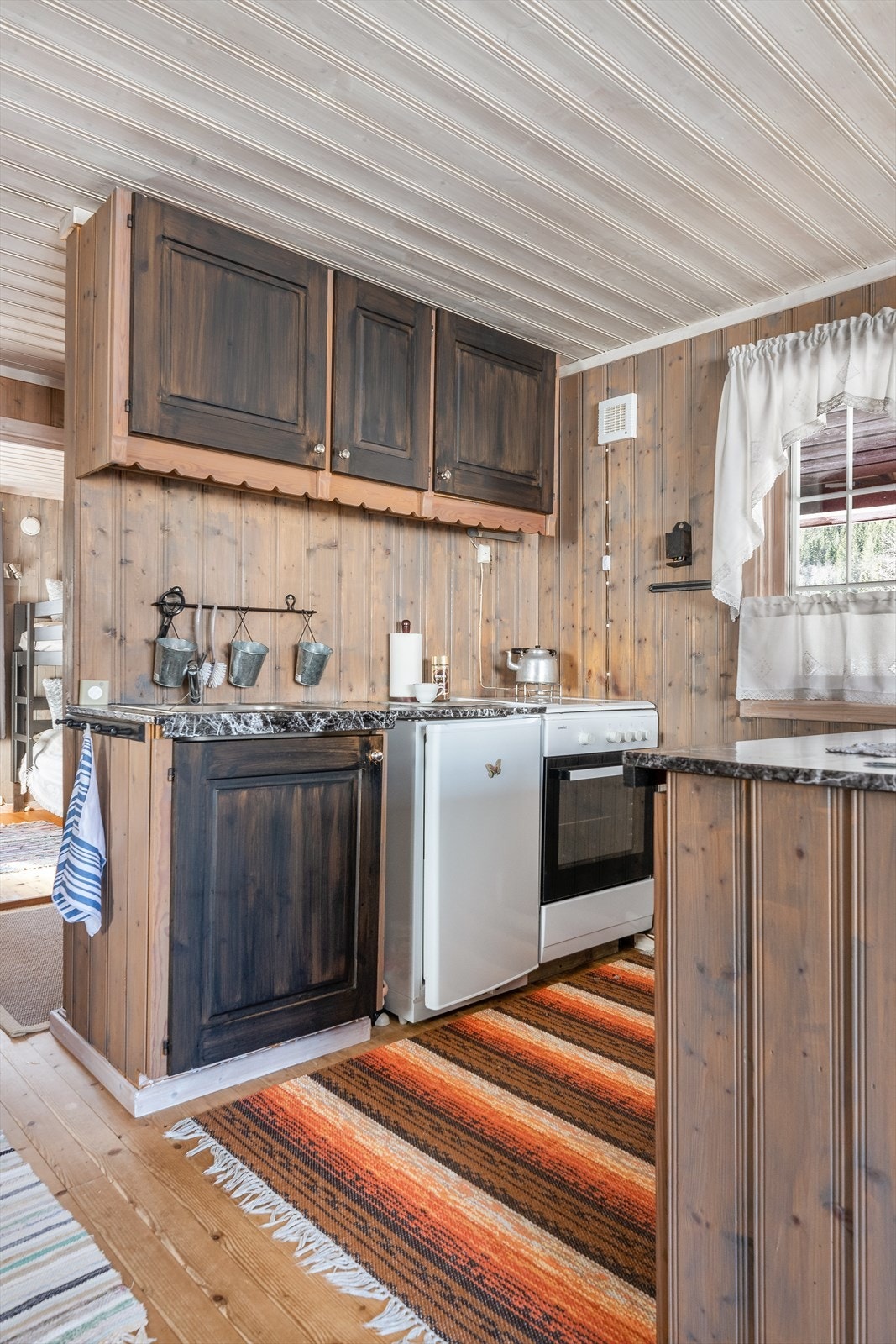 Skap og skuffer med god arbeidsflate over mot spisestuen. Avsatt plass til kjøleskap og komfyr som er styrt via gass. Det er installert gassvarsling både på kjøkken og i gang. Galleribilde