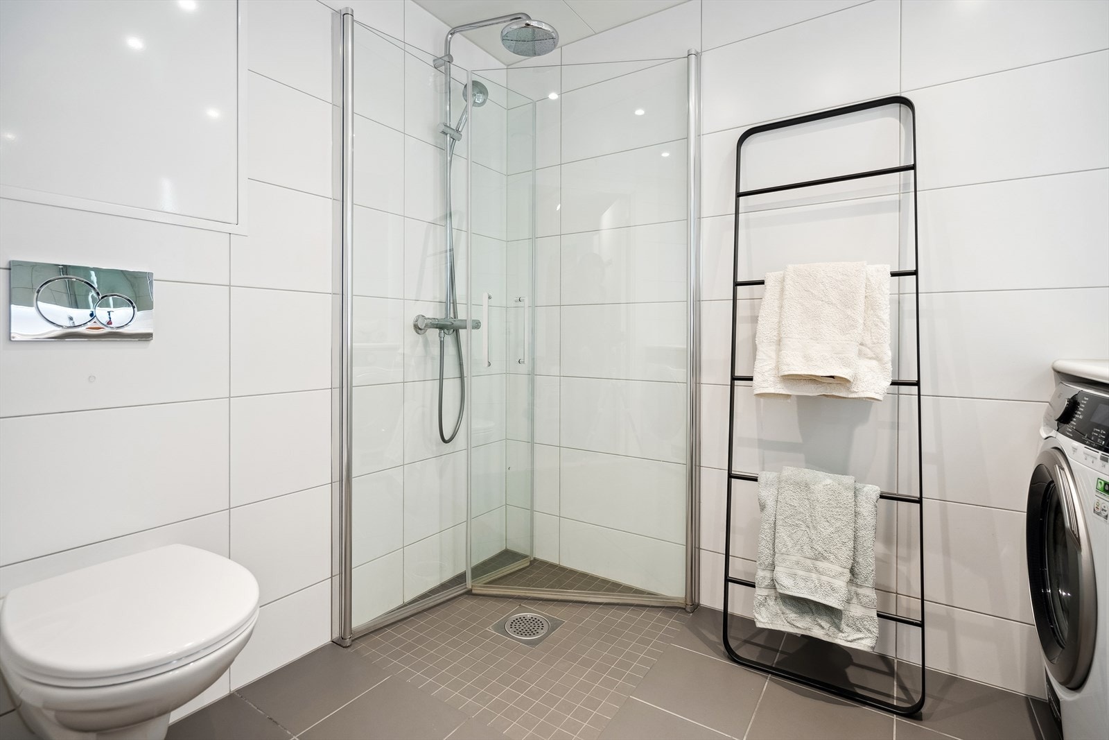 Badet er innredet med servantskap, servantbenkeplate, speil med lysarmatur, opplegg for vaskemaskin og tørketrommel, veggmontert klosett og dusjhjørne med regnfallsdusj. Galleribilde
