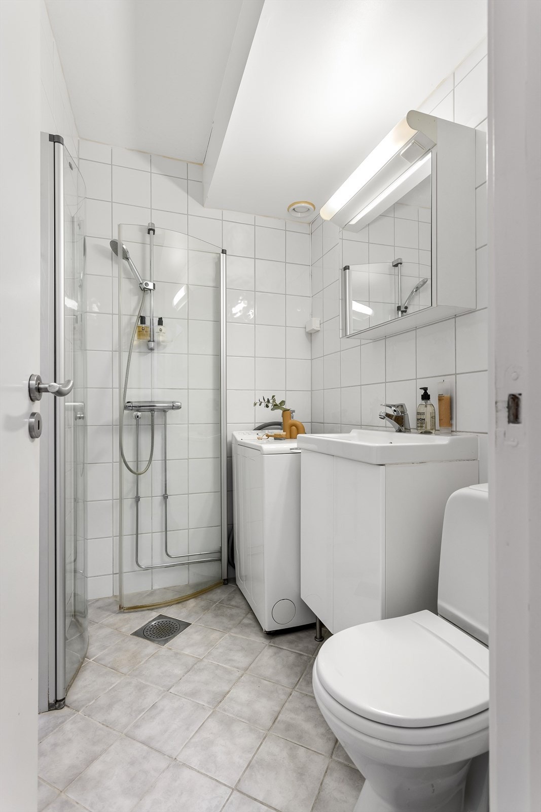 Dusjhjørne med vendbare, buede glassvegger, gulvstående toalett, servant med skap under, speilskap med lys og liten vaskemaskin (som kan medfølge). Galleribilde