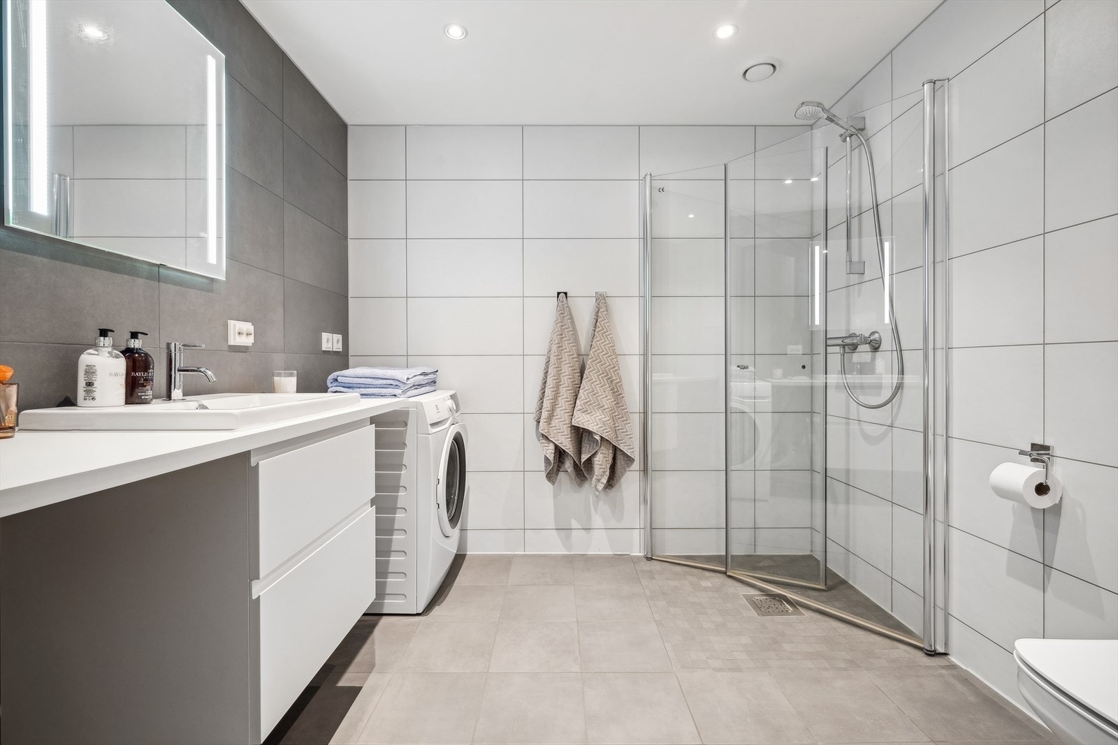 Pent og stort flislagt baderom innredet med downlights i taket. Innredningen består av servantinnredning med speil, skuffer, vegghengt klosett og praktisk dusjhjørne med innfellbare dører. Galleribilde