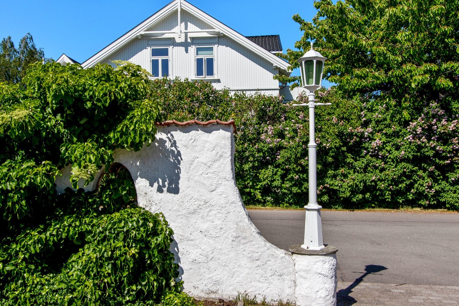 Mot Teglverksveien er det hvitmalt mur og innkjøring mellom to stolper med utelamper. Galleribilde