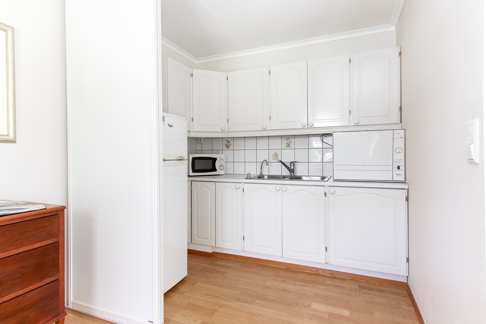 Kjøkkeninnredningen er enkel med benkeskap, overskap og dobbel oppvaskkum i en hvit laminatbenkeplate. Galleribilde