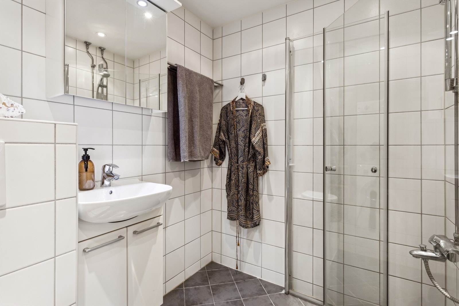 Baderomsinnredning med heldekkende servant, vegghengt toalett, speilskap og dusjhjørne med innfellbare glassdører. Galleribilde