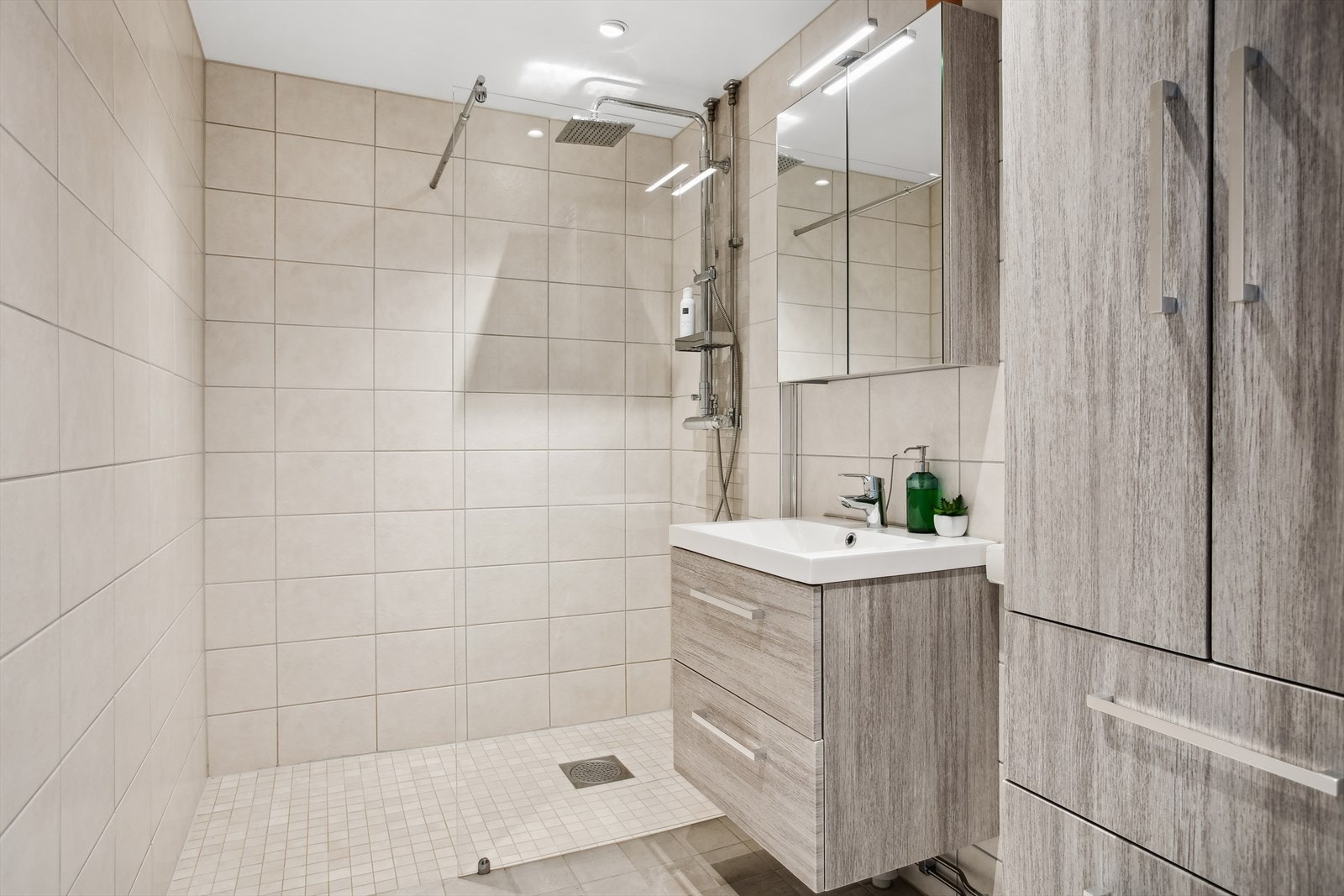 Innredet med stor dusjhjørne med fastmontert glassvegg, termostatstyrt dusjbatteri, regndusj/hånddusj. Servantinnredning med laminerte fronter i i imitert treverk, servantskap med skuffer, speilskap med LED belysning og vegghengt høyskap i lik utfør. Galleribilde