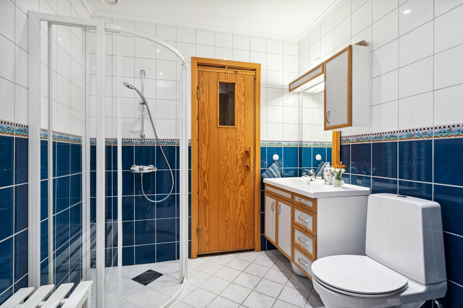 Bad 2 i 1.etasje har adkomst fra hall. Flislagte overflater, gulvvarme, innredning, dusjhjørne og toalett. Her er det dør inn til badstuen Galleribilde