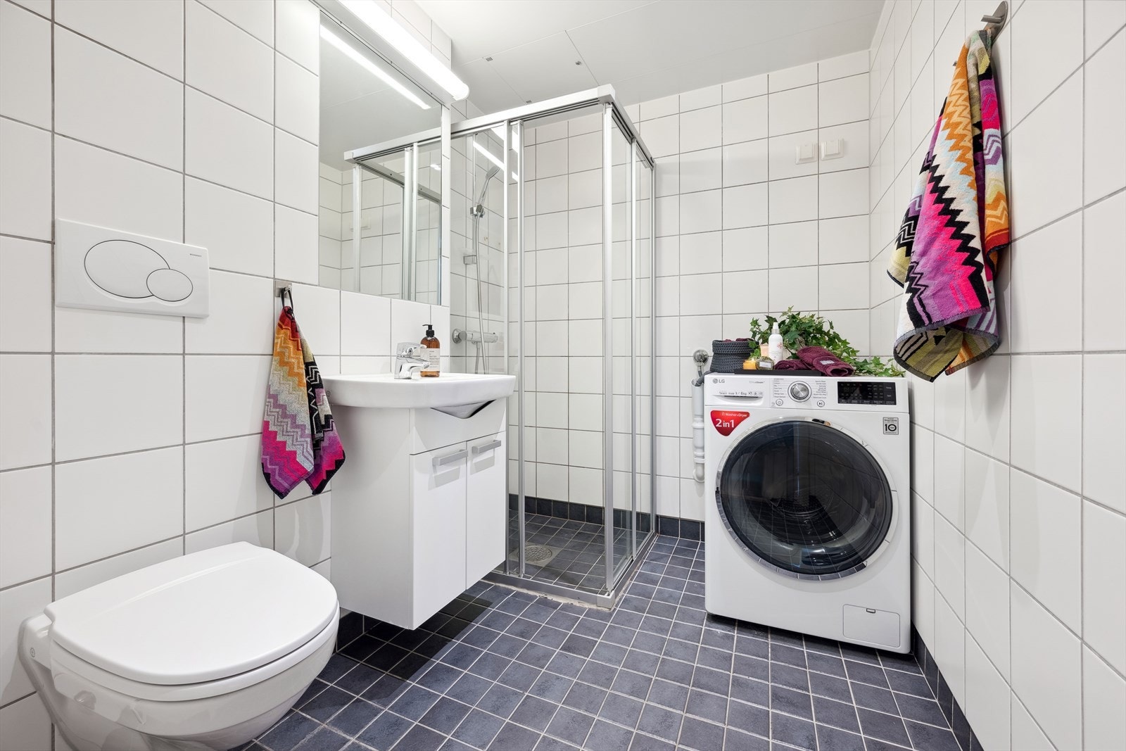 Badet er innredet med servant på underskap, dusjhjørne, vegghengt toalett og opplegg for vaskemaskin. Galleribilde